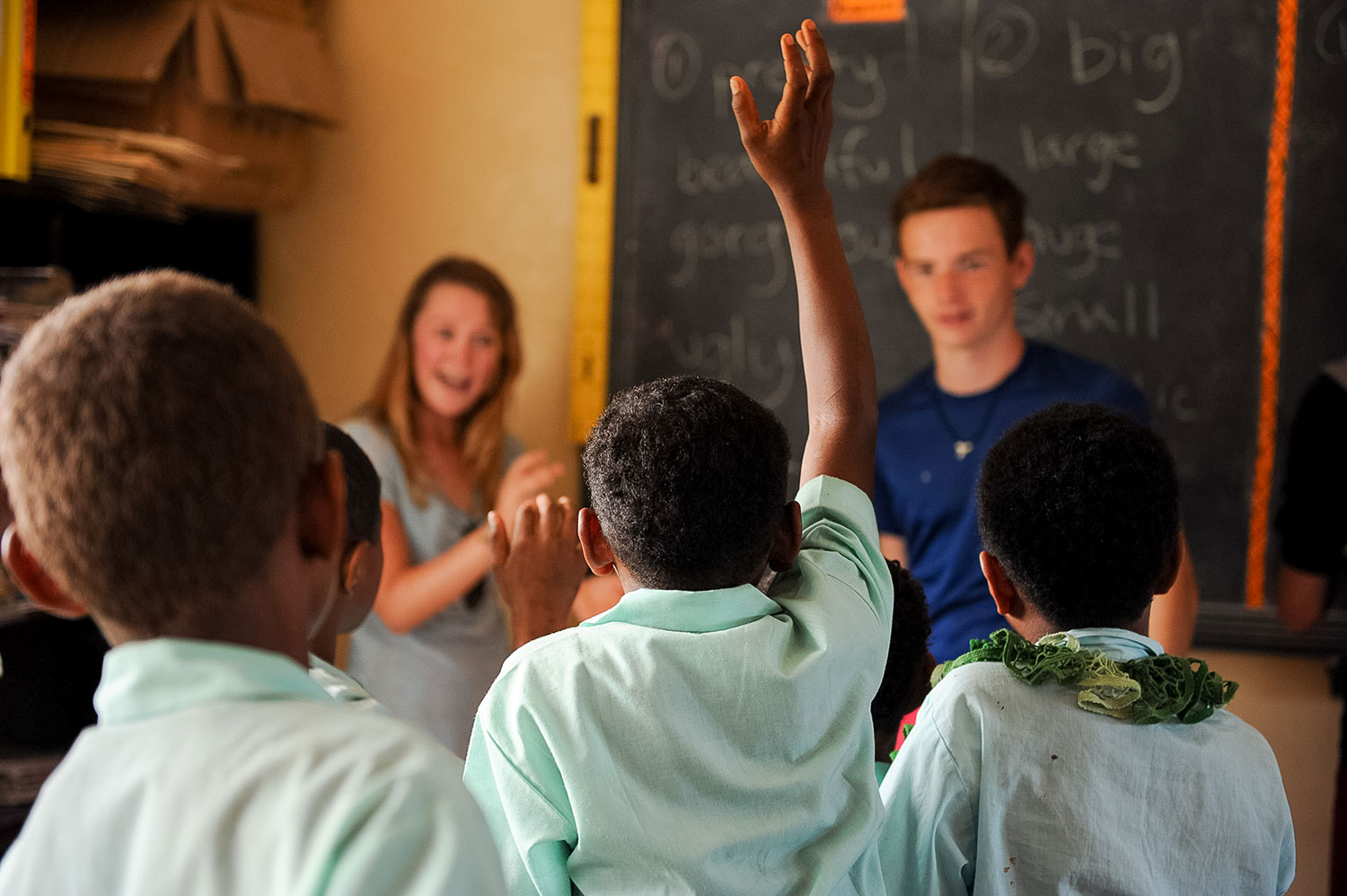 A student raises a hand during a classroom lesson with a chalkboard behind