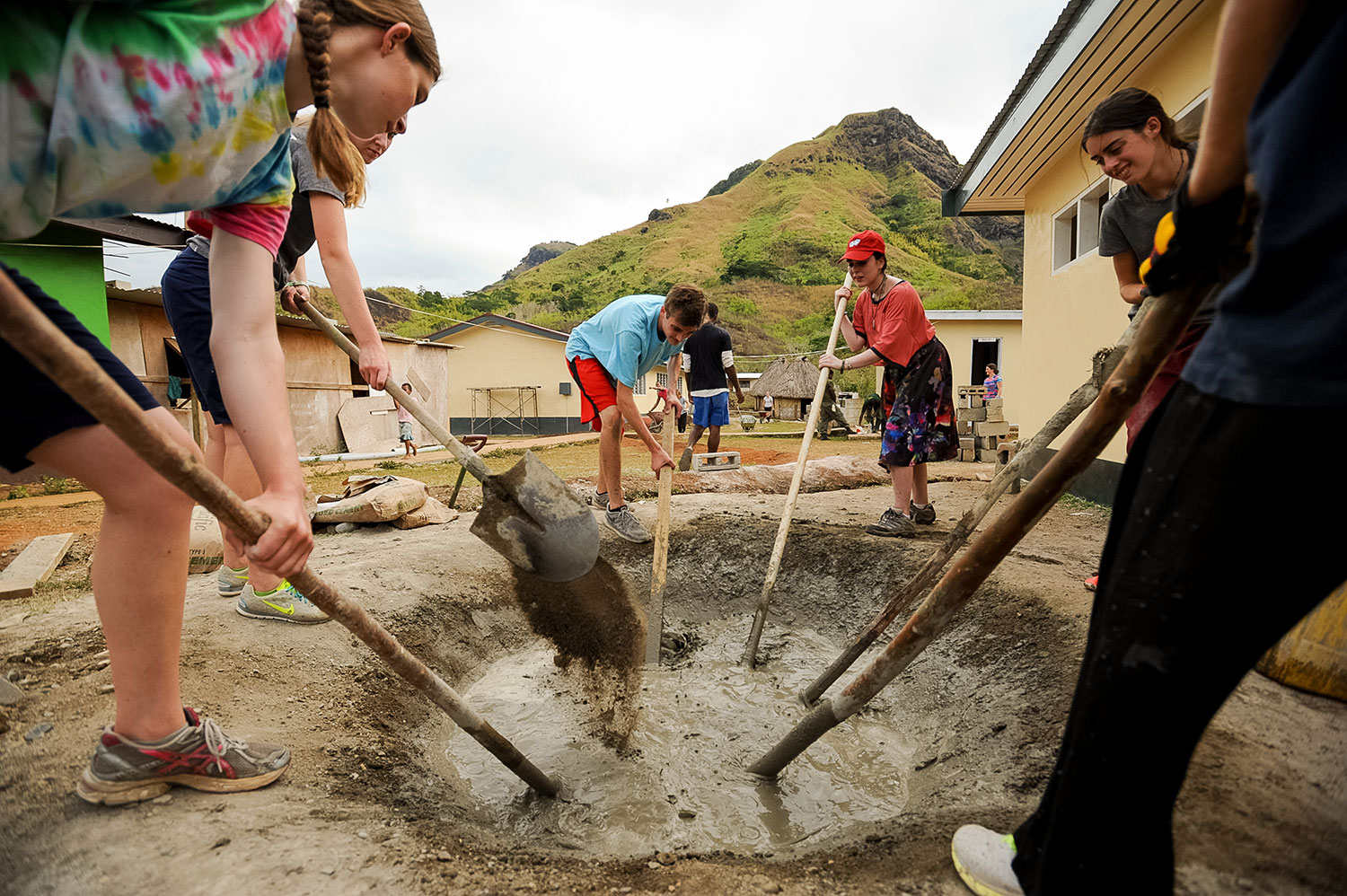 Students mix and pour concrete into a mold with hills rising behind a village in Fiji