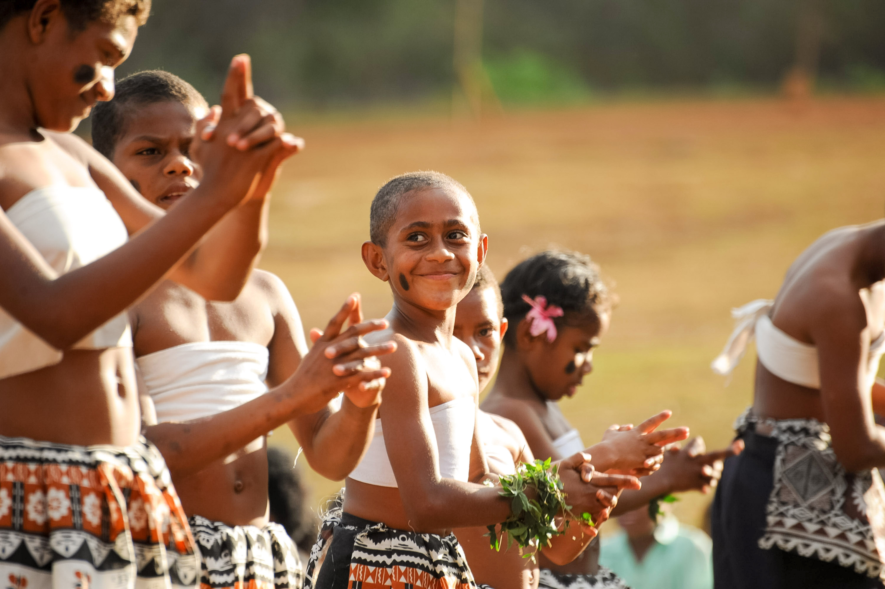 A young Fijian dancer smiles mid-performance in traditional dress with others clapping alongside