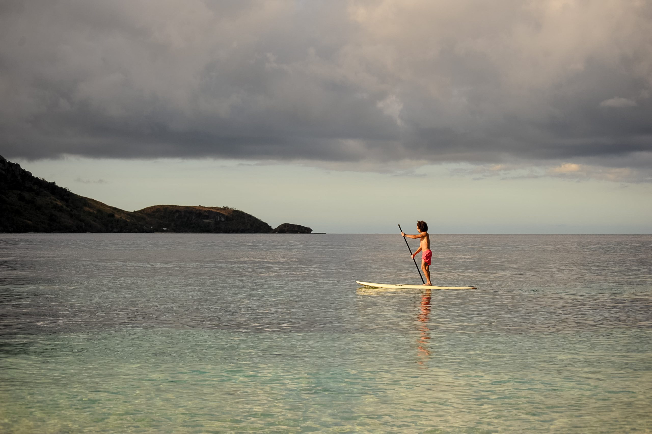 A student paddleboards on calm water with hills in the distance