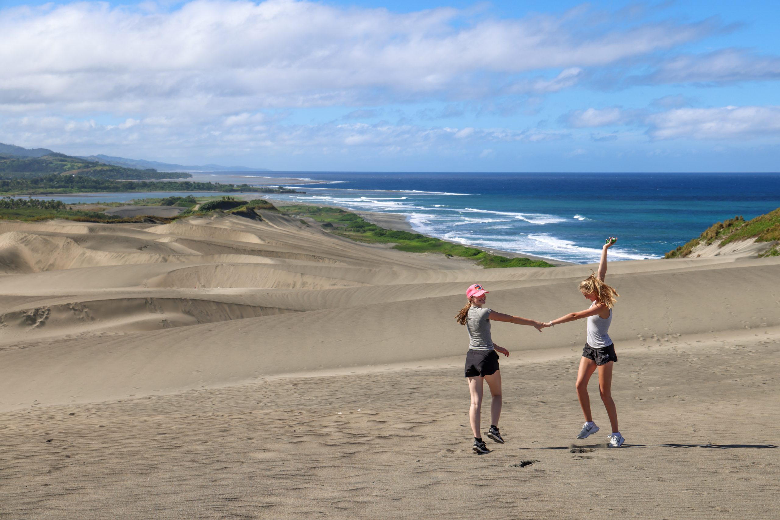 Students hold hands while jumping on a sand dune overlooking the coastline