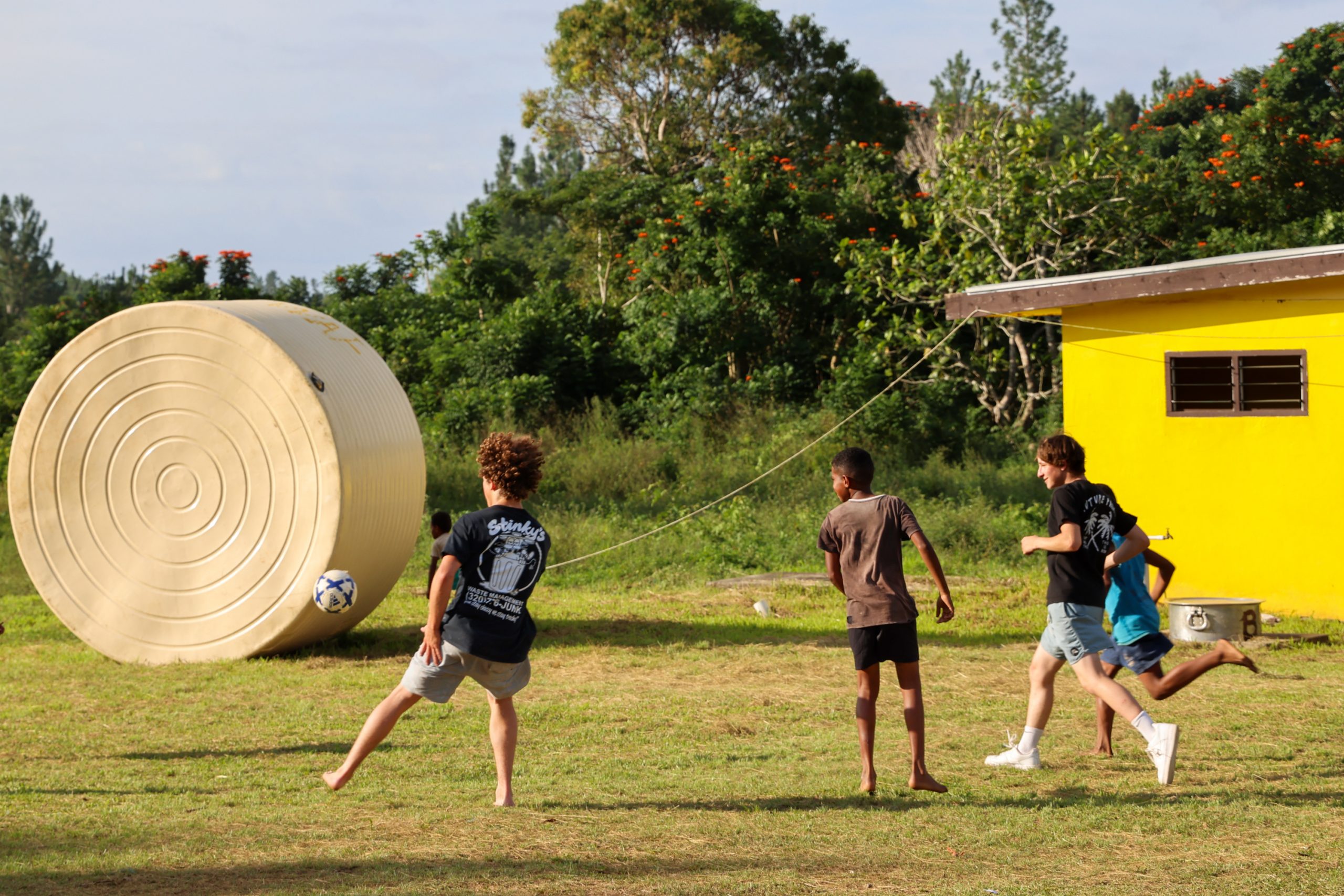 Students play soccer on a grassy field near a large water tank and yellow building