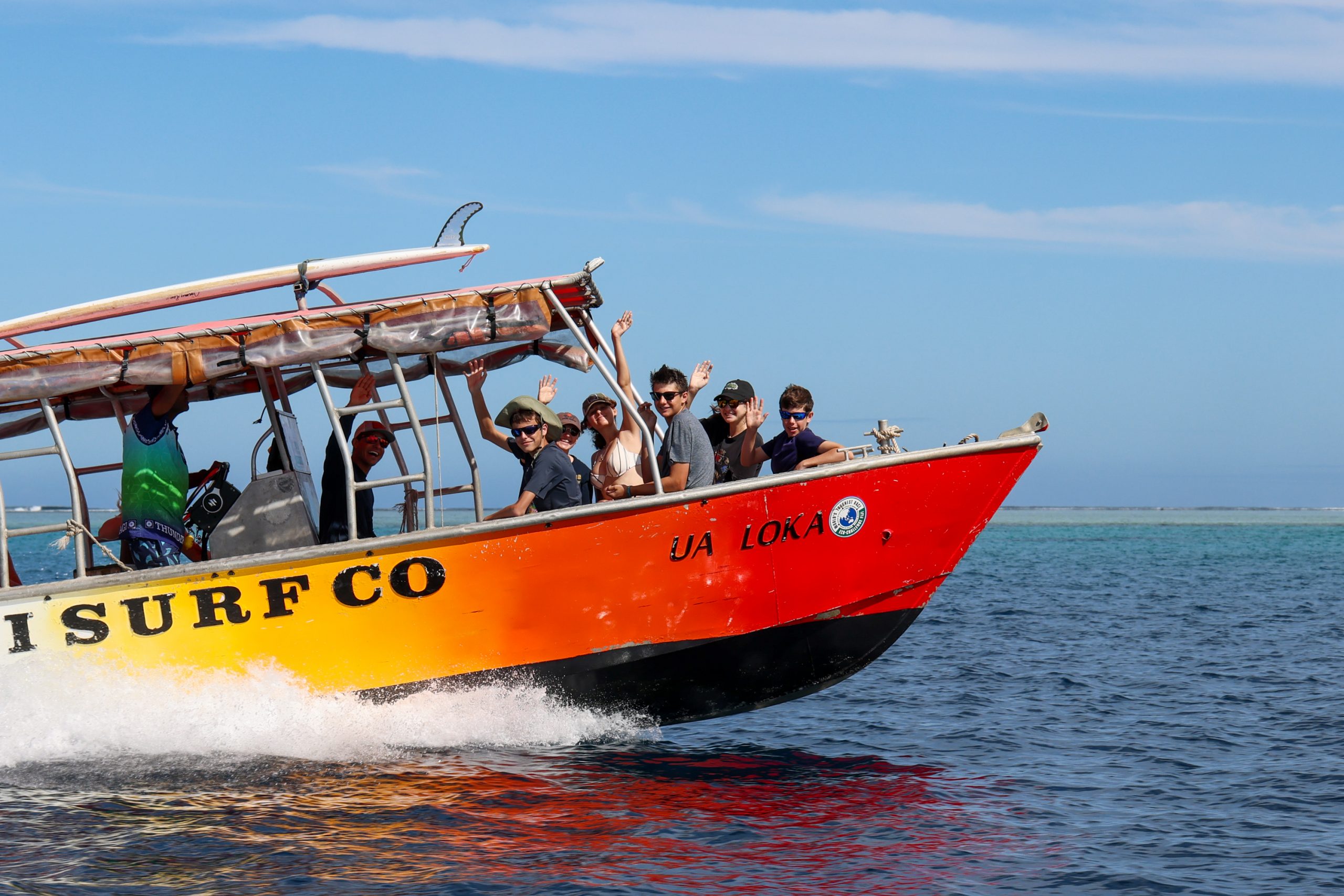 Students wave from a surf boat speeding across turquoise water in Fiji