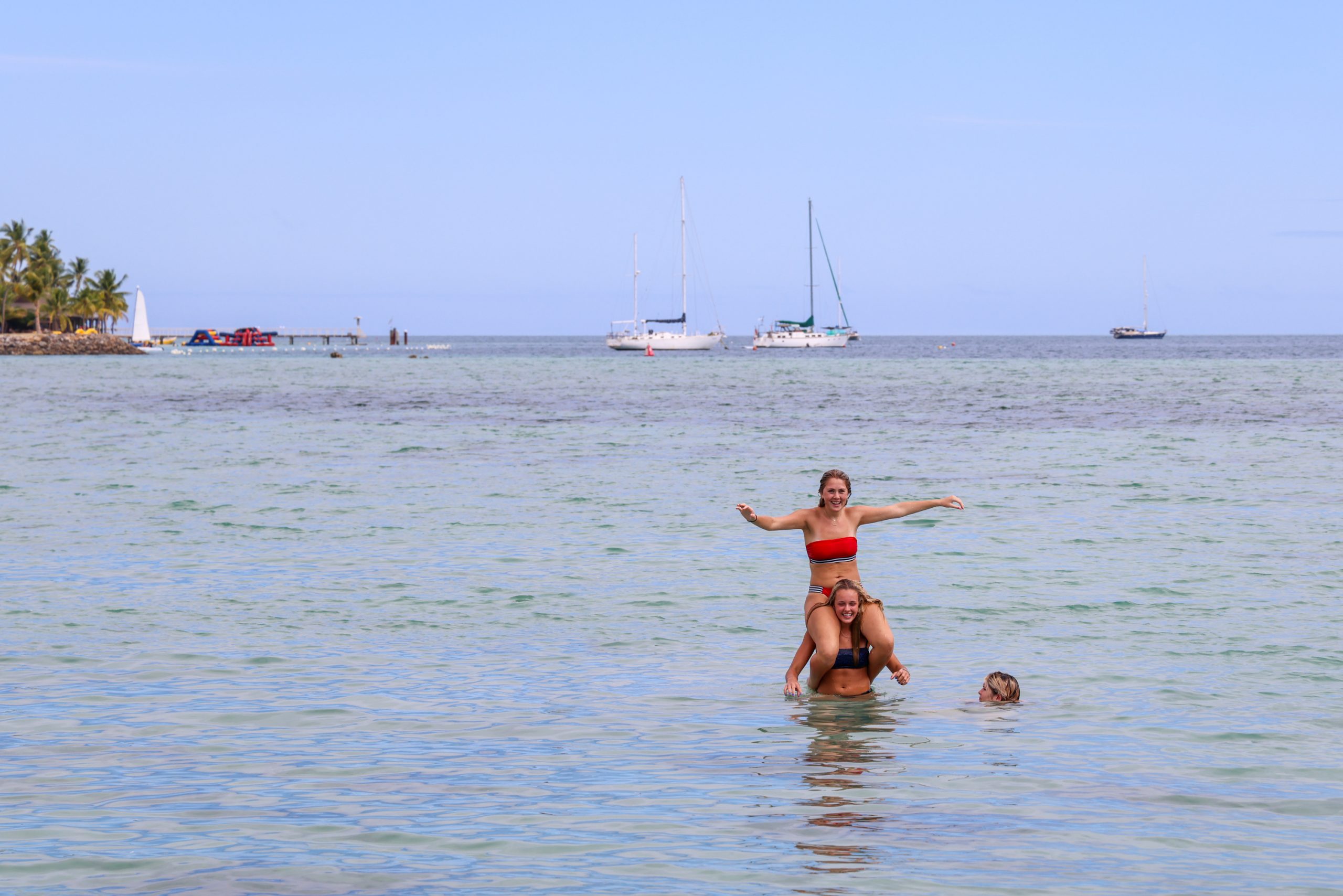 Students play in shallow water with sailboats anchored offshore
