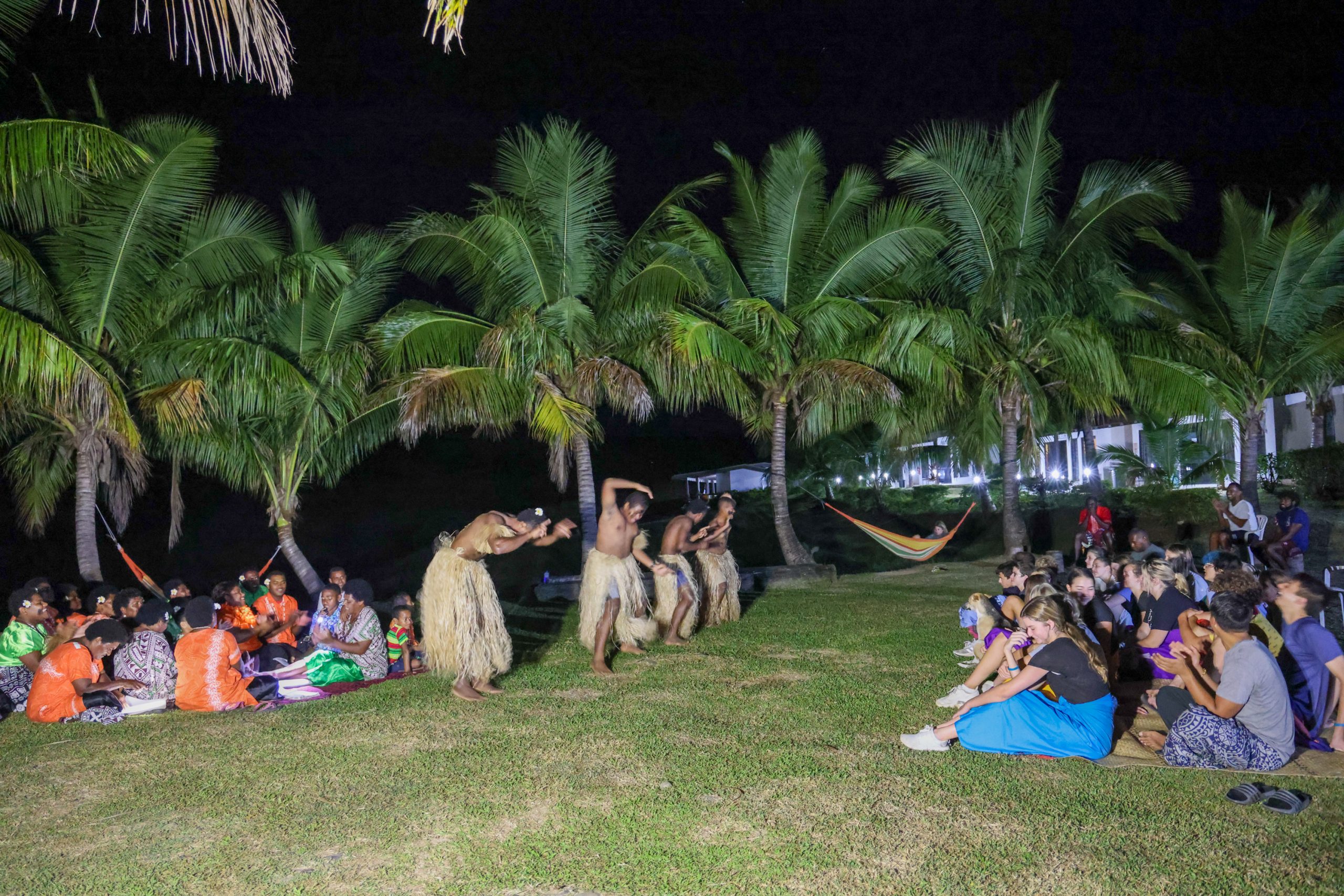 Traditional dancers perform under palm trees at night as students watch from the grass