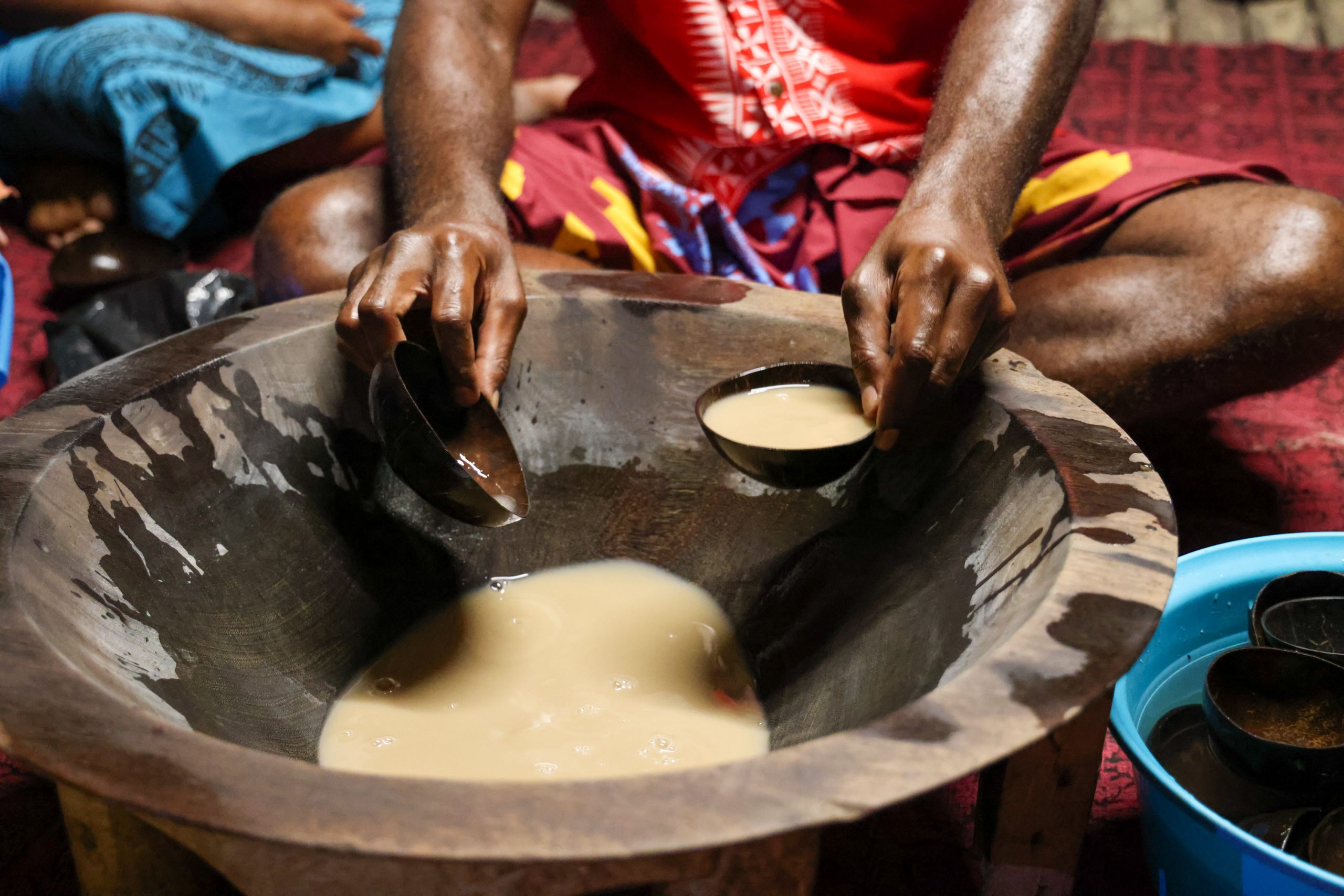 Hands ladle kava from a wooden bowl into small cups during a traditional ceremony