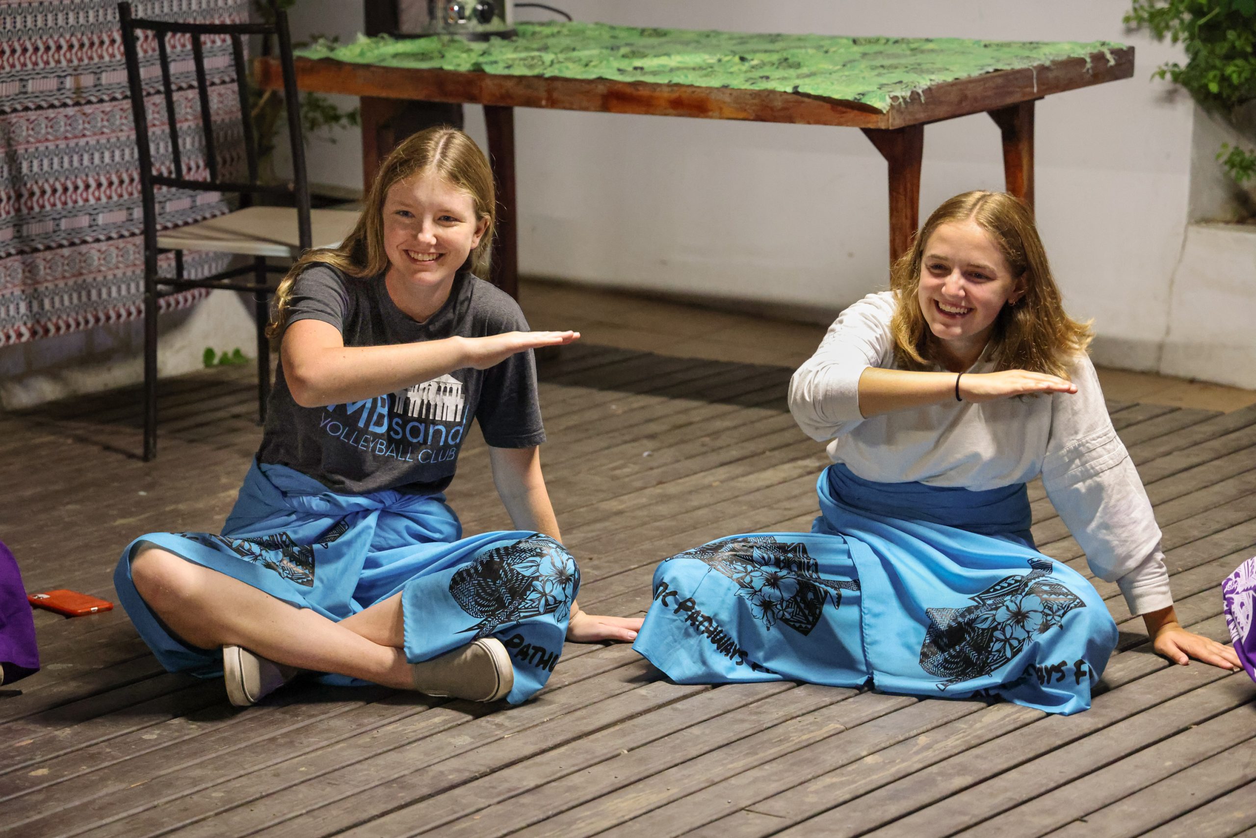 Students in traditional Fijian sulu skirts pose mid-dance on a wooden deck