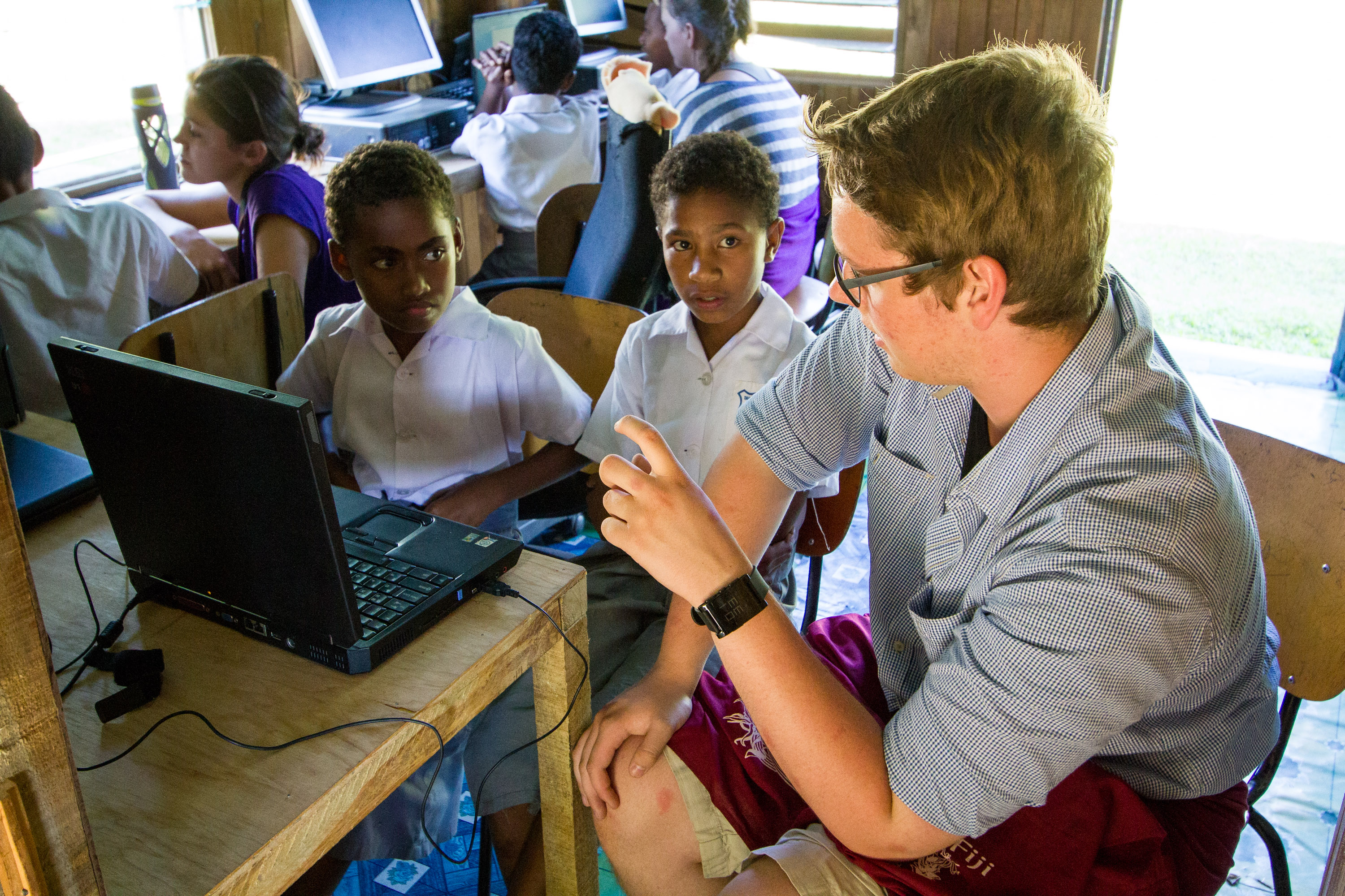 A volunteer shows local schoolchildren something on a laptop in a classroom