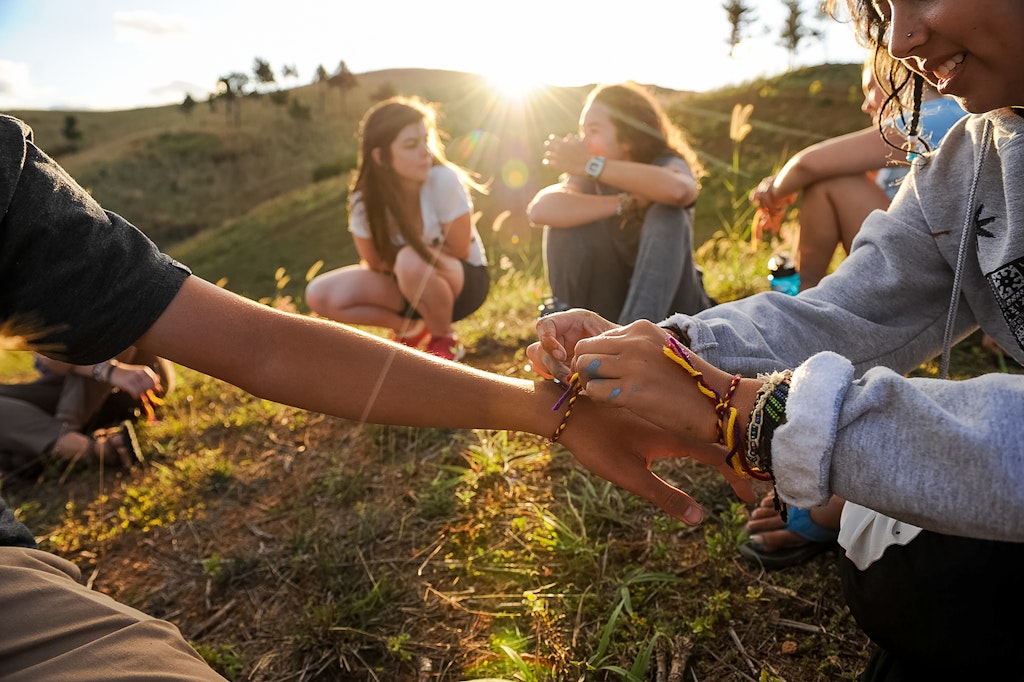 Friends are sitting on the grass, exchanging bracelets as the sunlight shines into the camera.