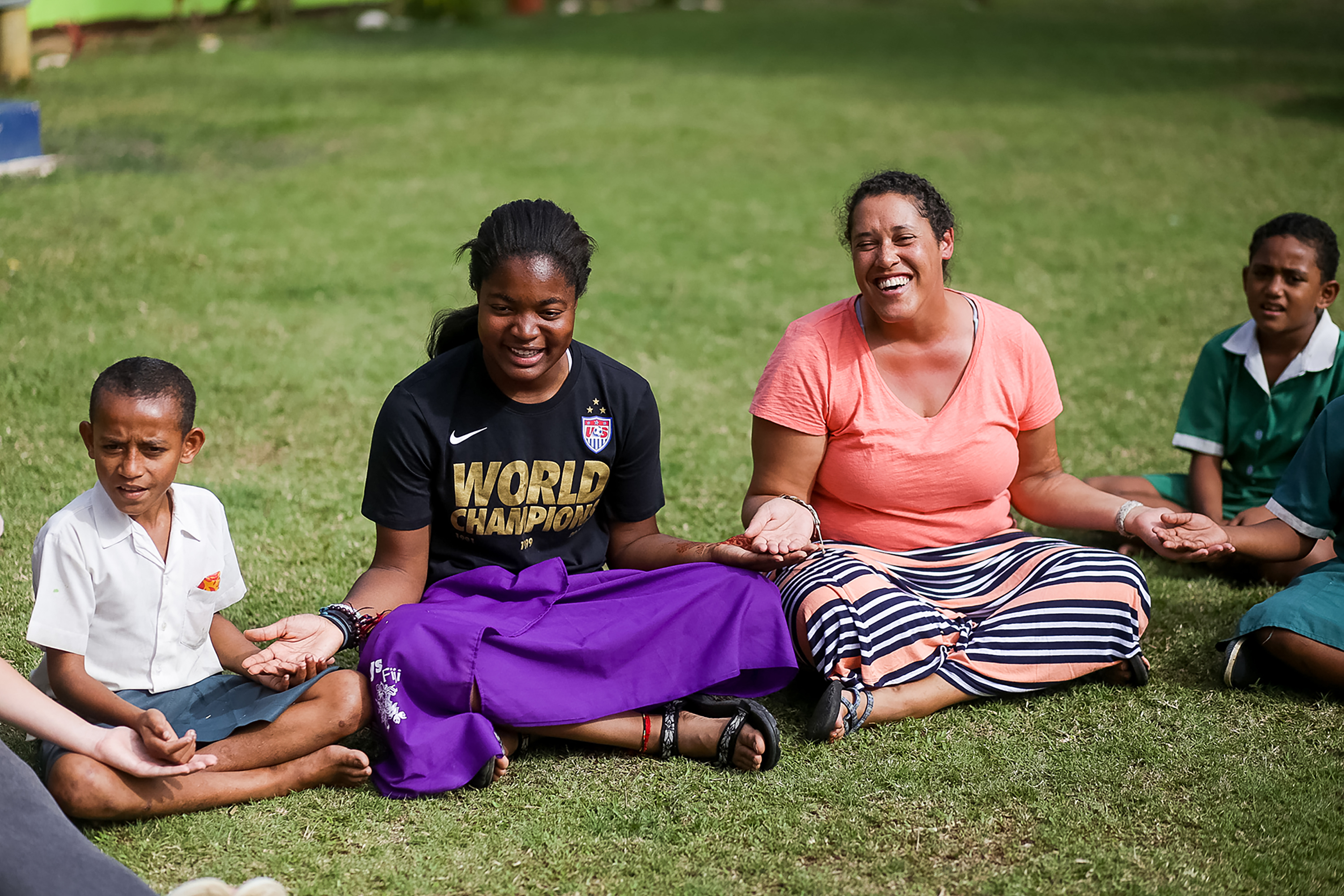 Students and local children sit cross-legged on grass, laughing together