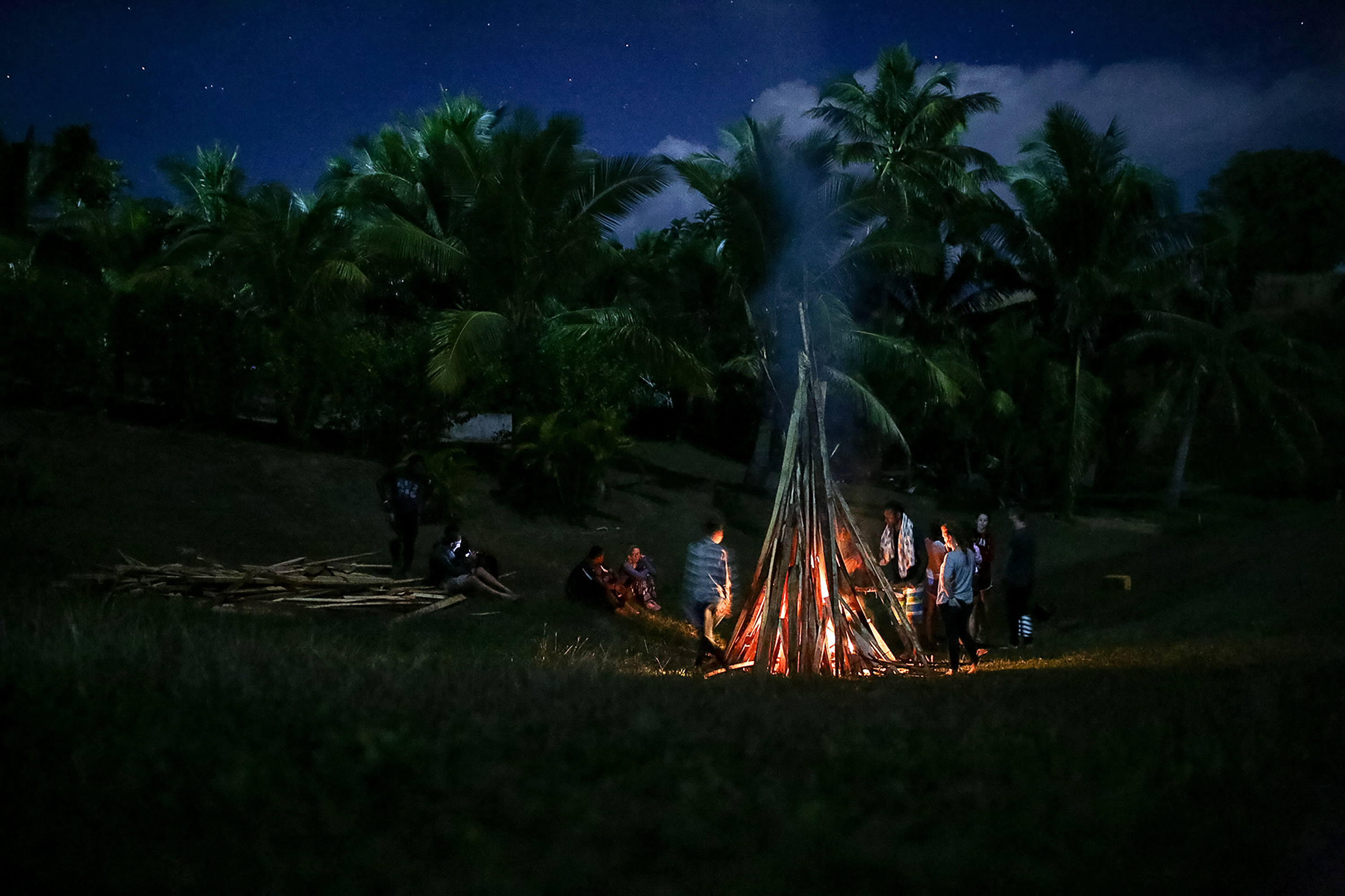 Students gather around a large bonfire at night beneath palm trees