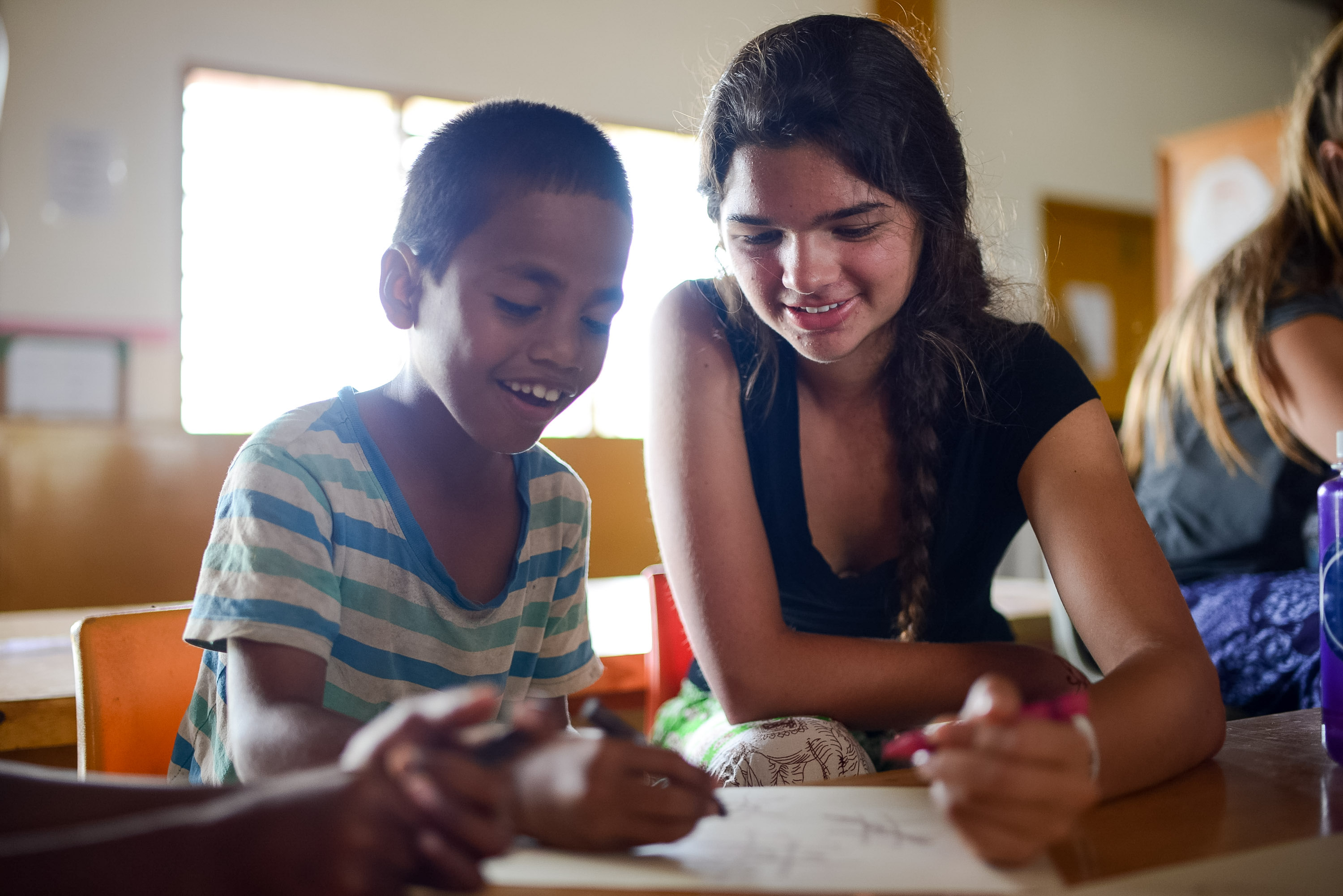 A student helps a young boy with writing at a classroom desk