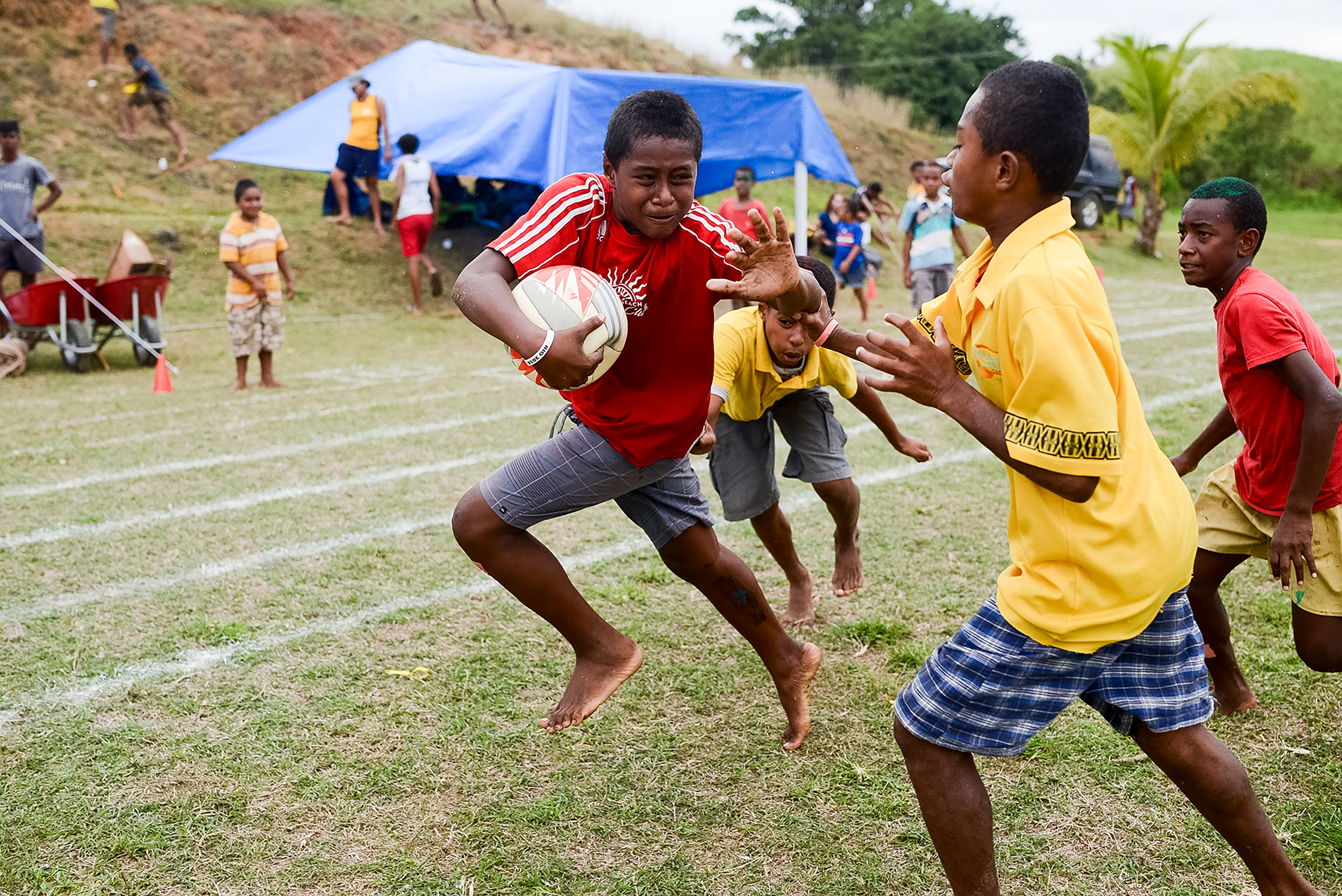 A boy runs with a rugby ball while others give chase on a grass field in Fiji