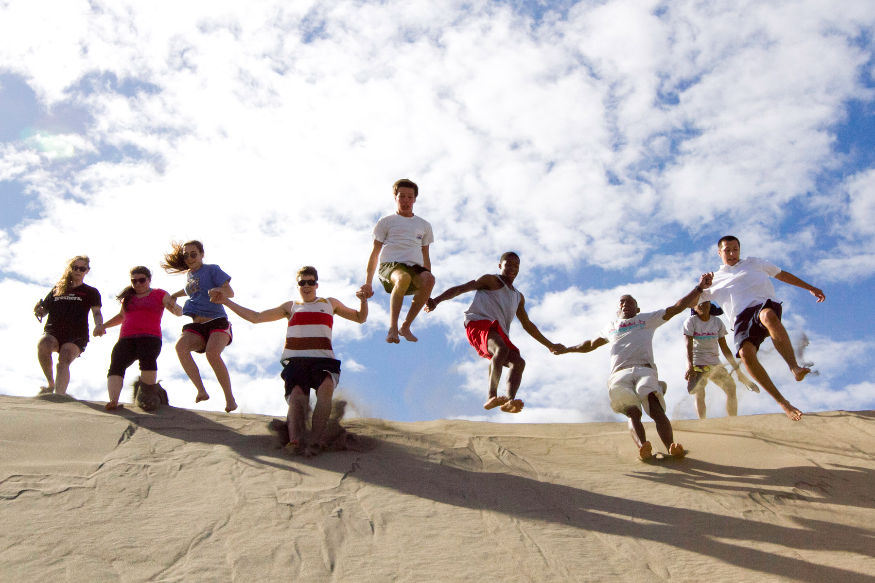 Students hold hands and jump together off a sand dune