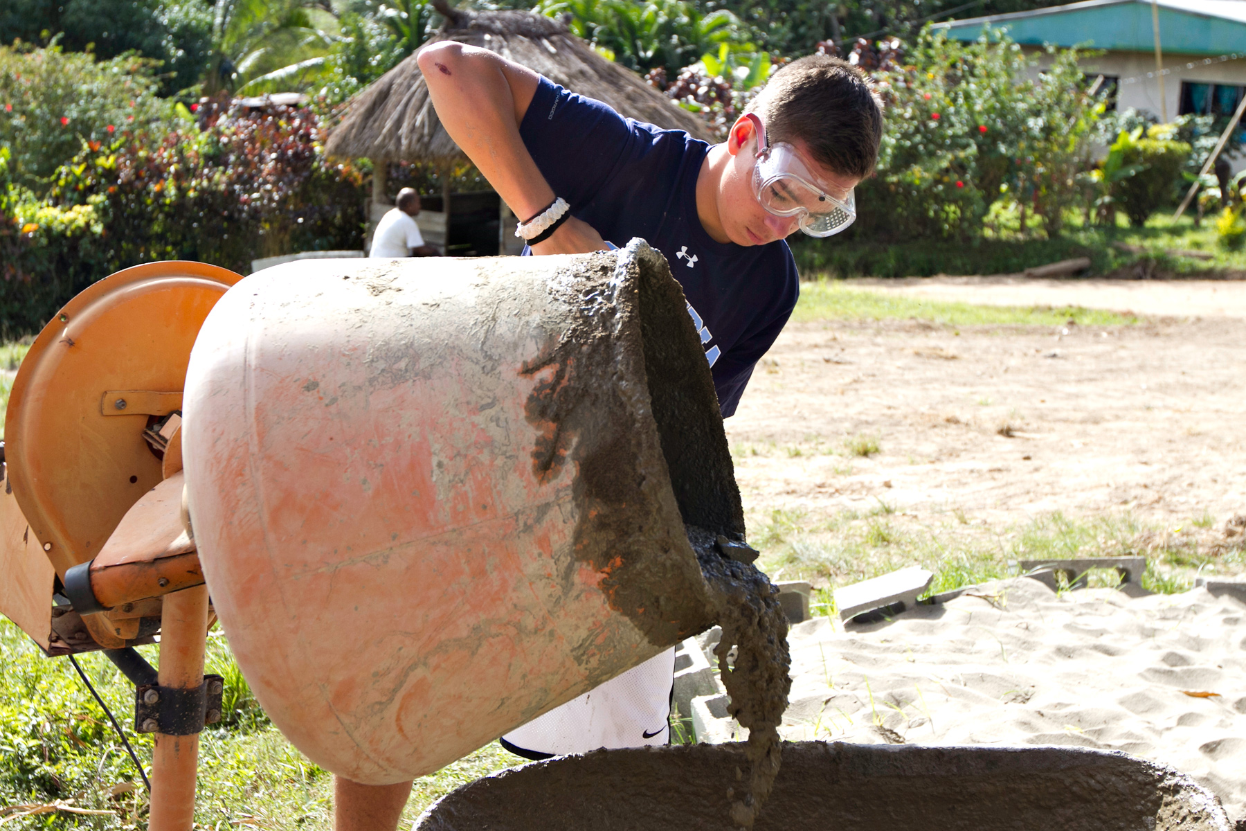 A student wearing safety goggles pours concrete from a mixing drum at a construction site