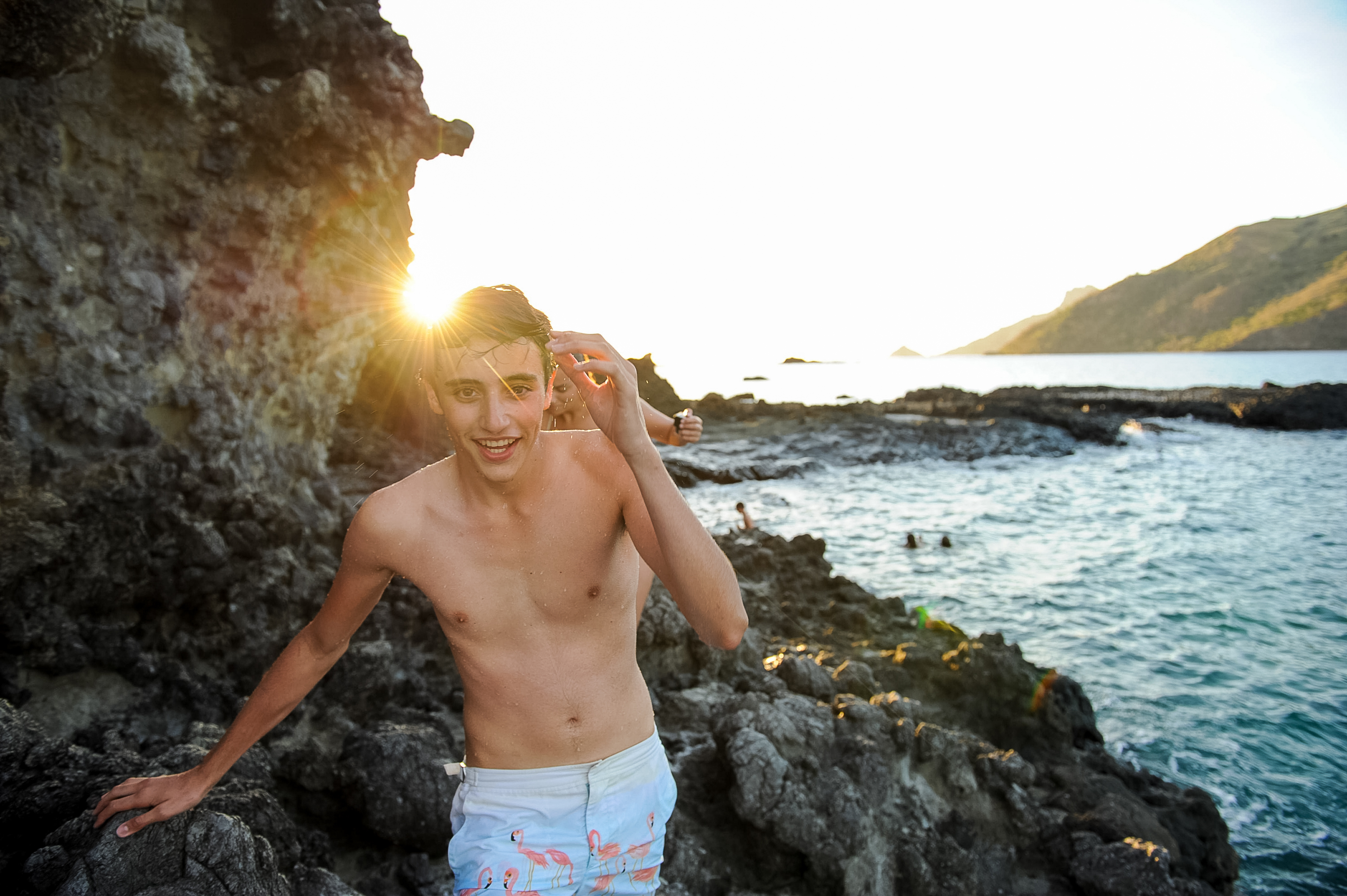On a school trip, a student stands on volcanic rocks along the coastline at sunset