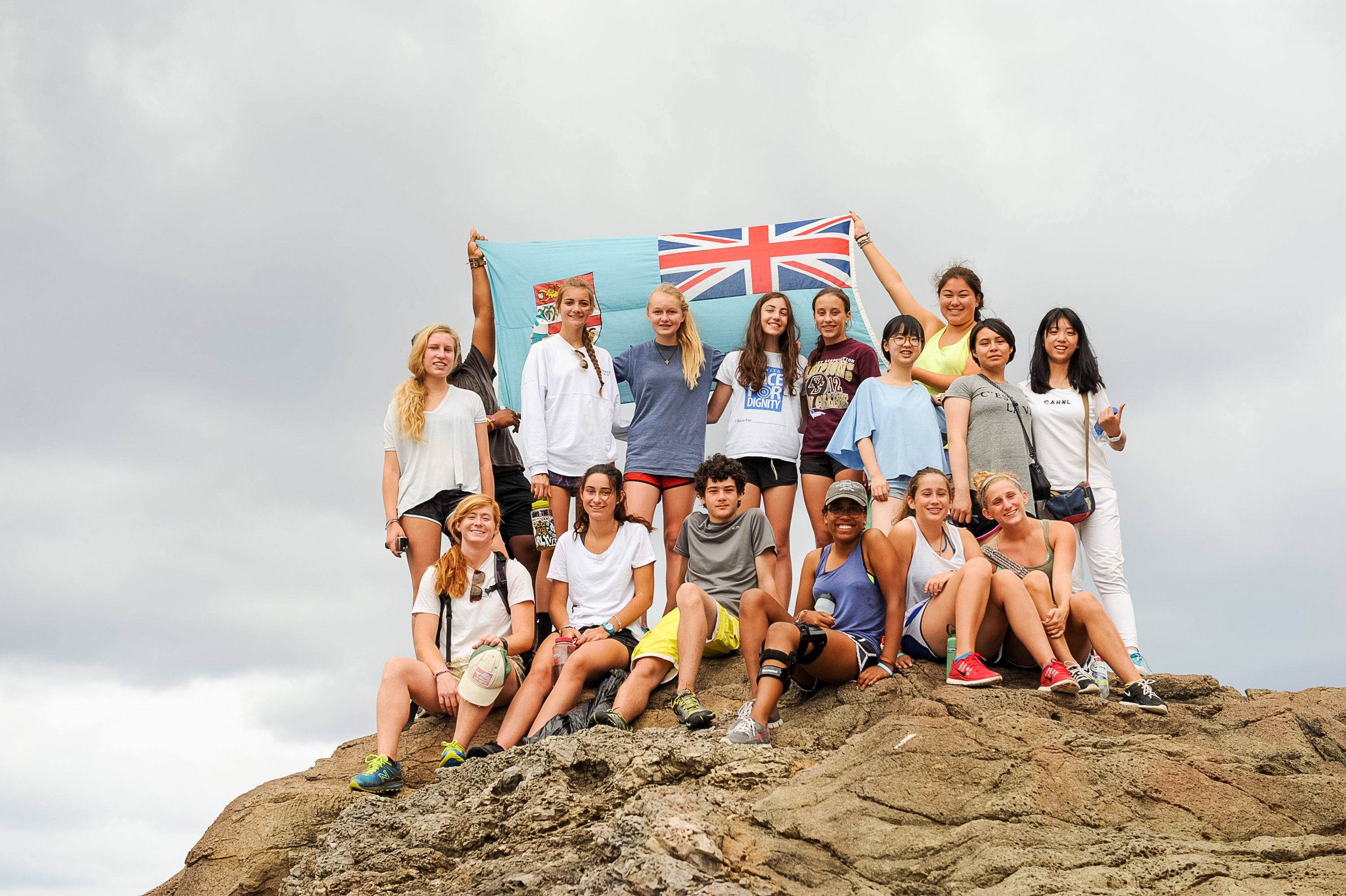 Students pose atop a rocky summit holding a Fijian flag under cloudy skies