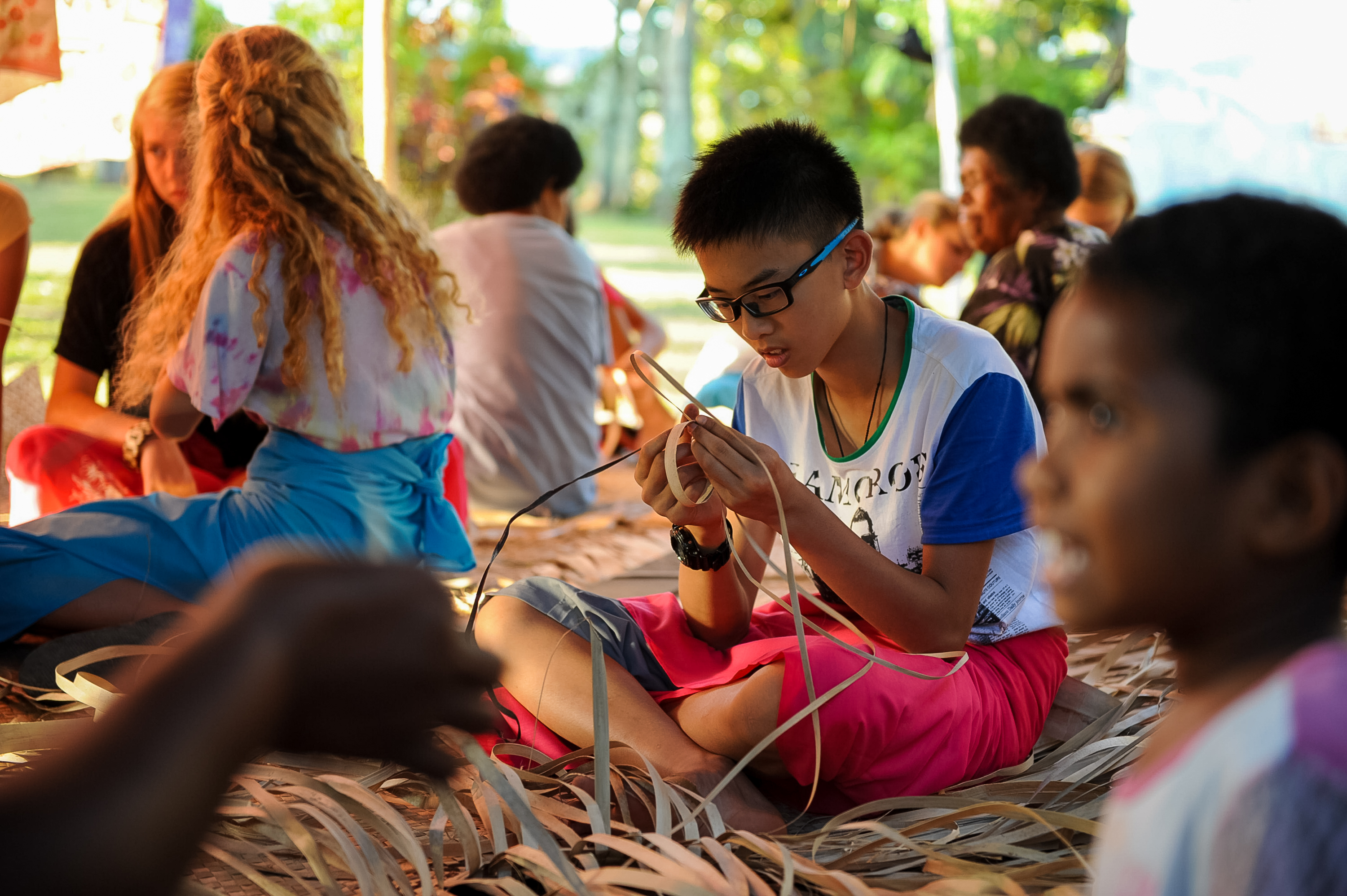 A student weaves palm fronds while sitting on a woven mat with local community members