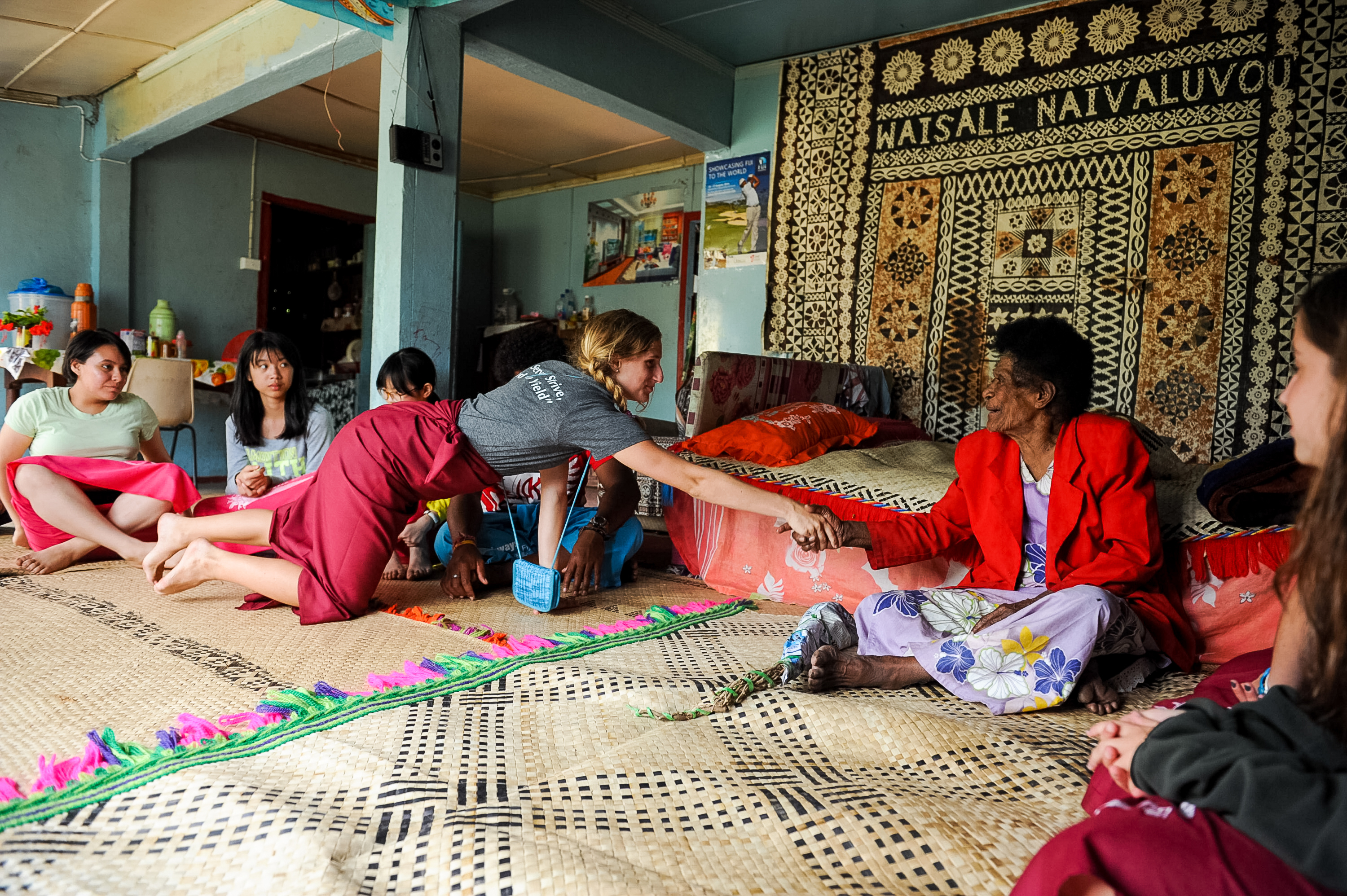 Students sit on woven mats exchanging a garland with a village elder in a decorated Fijian hall