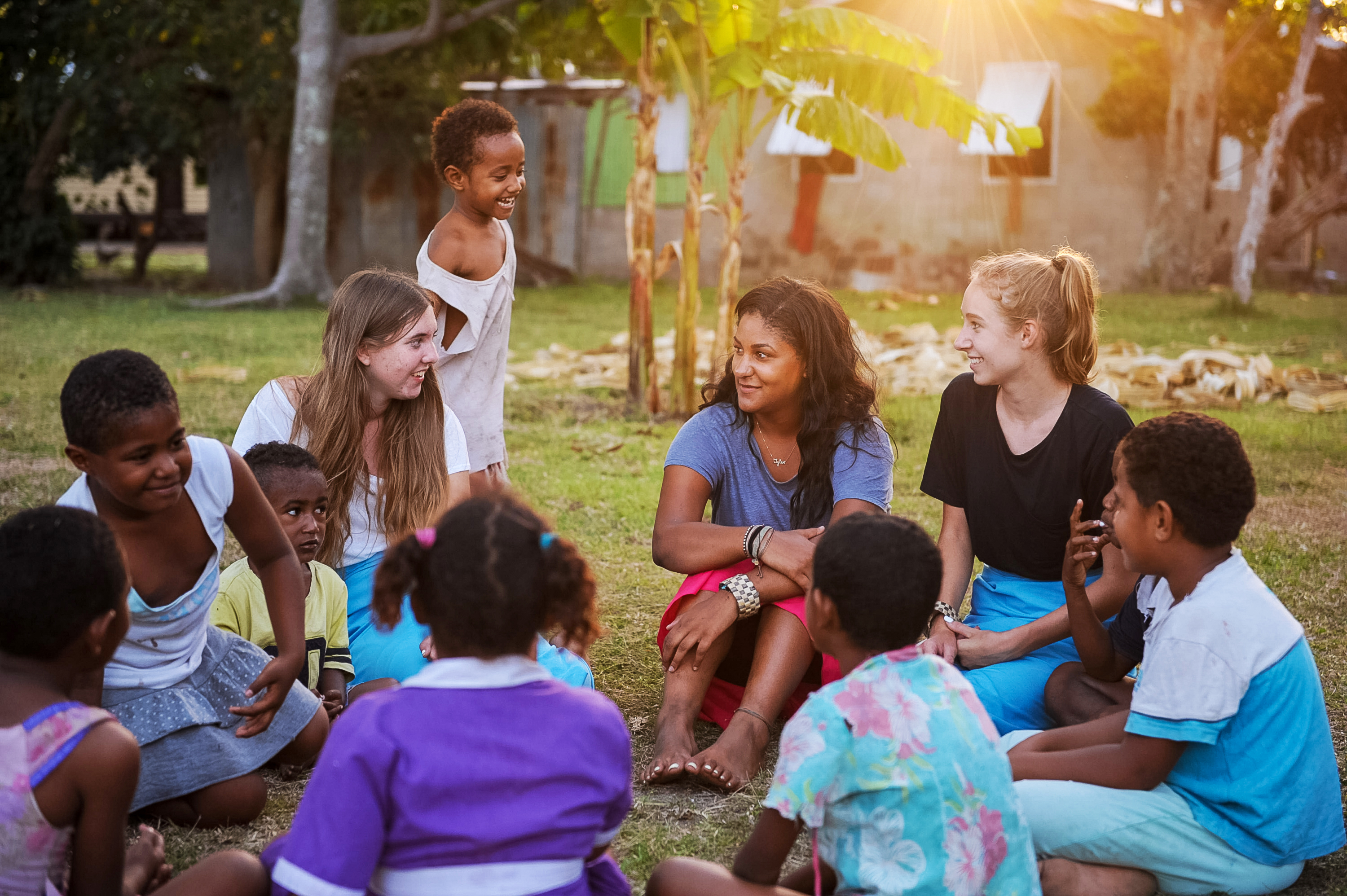 Students sit in a circle with local children in a grassy village yard