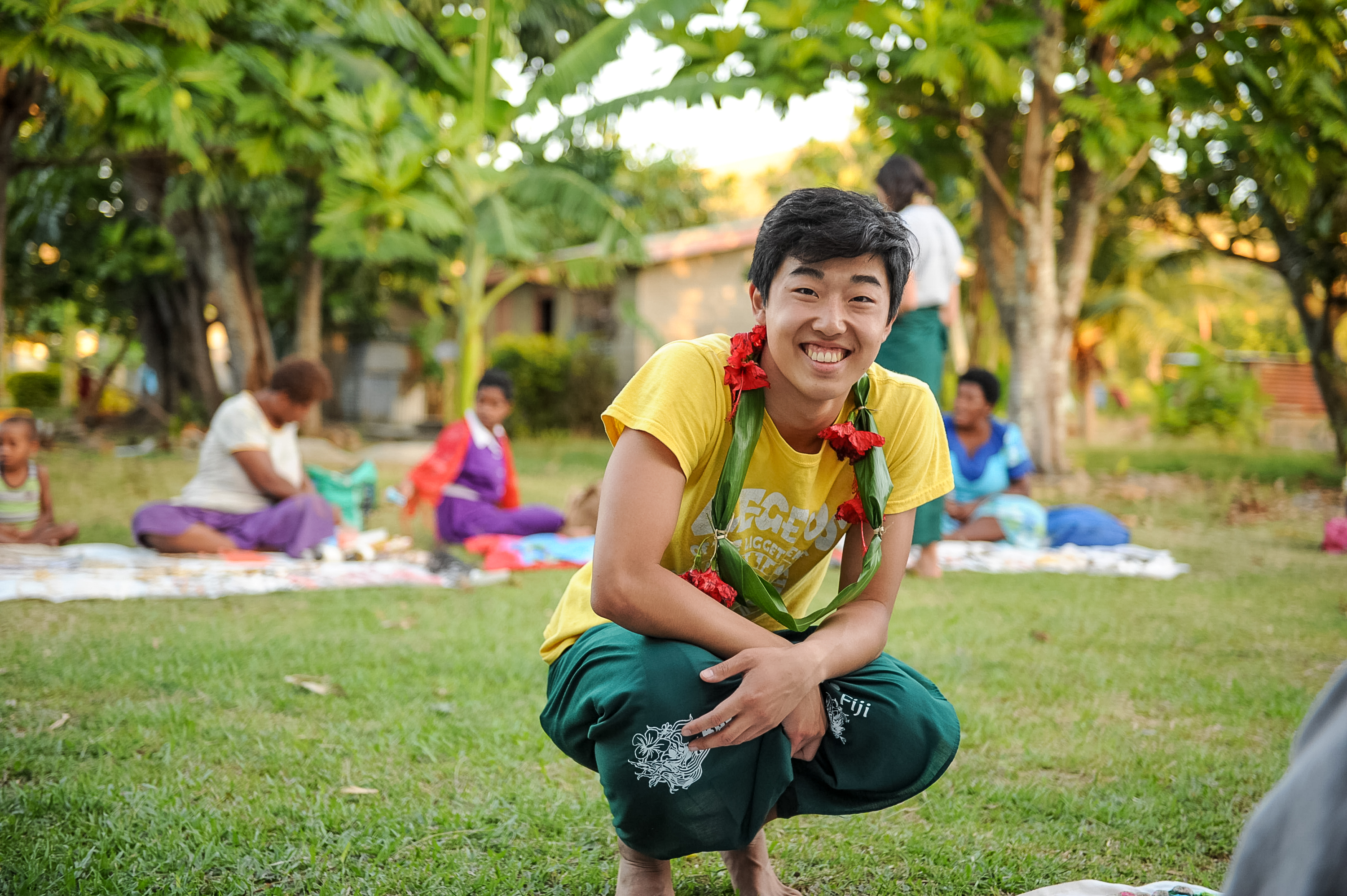 A student wearing a floral lei crouches smiling on the grass with local community members seated nearby