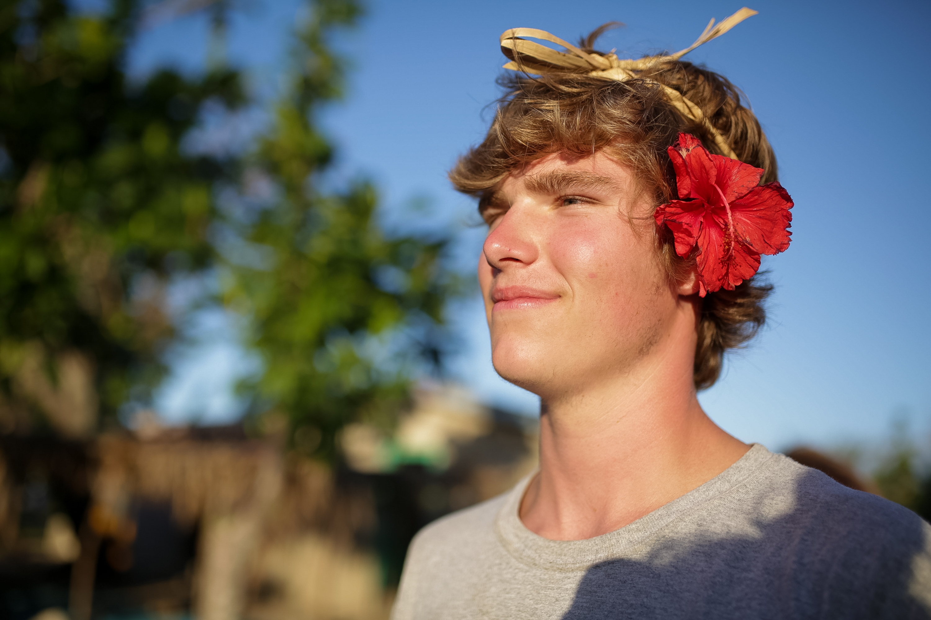 A student wears a red hibiscus flower and a woven headband, smiling in the sunlight