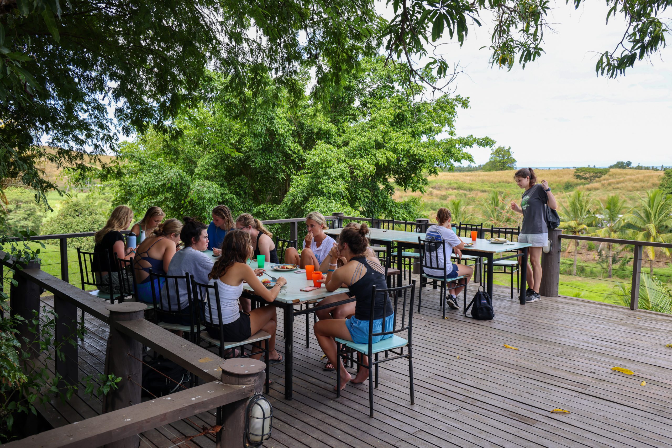 Students sit at outdoor tables on a wooden deck overlooking green hills