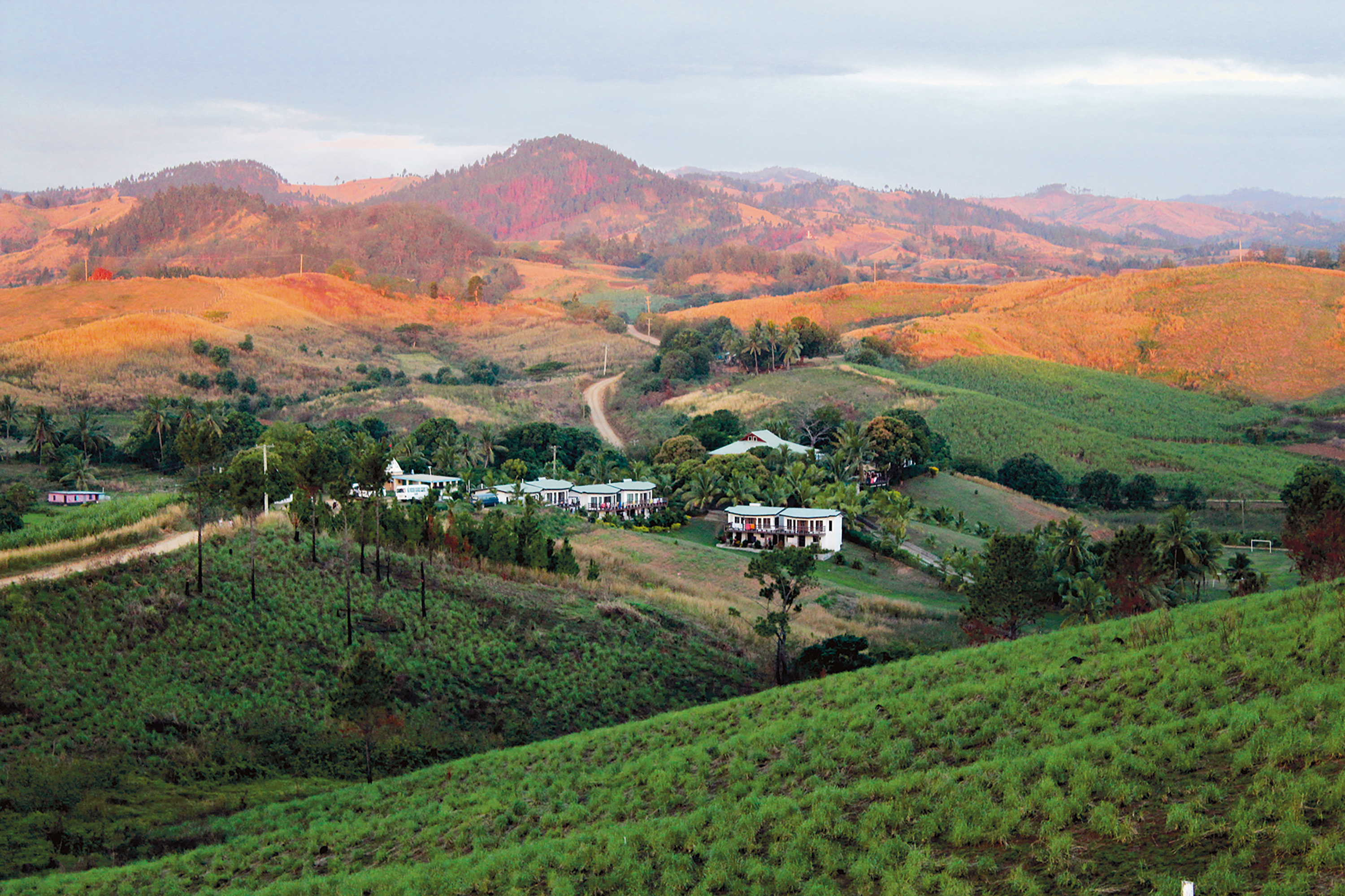 A small village nestled among rolling green hills with a dirt road winding toward distant mountains