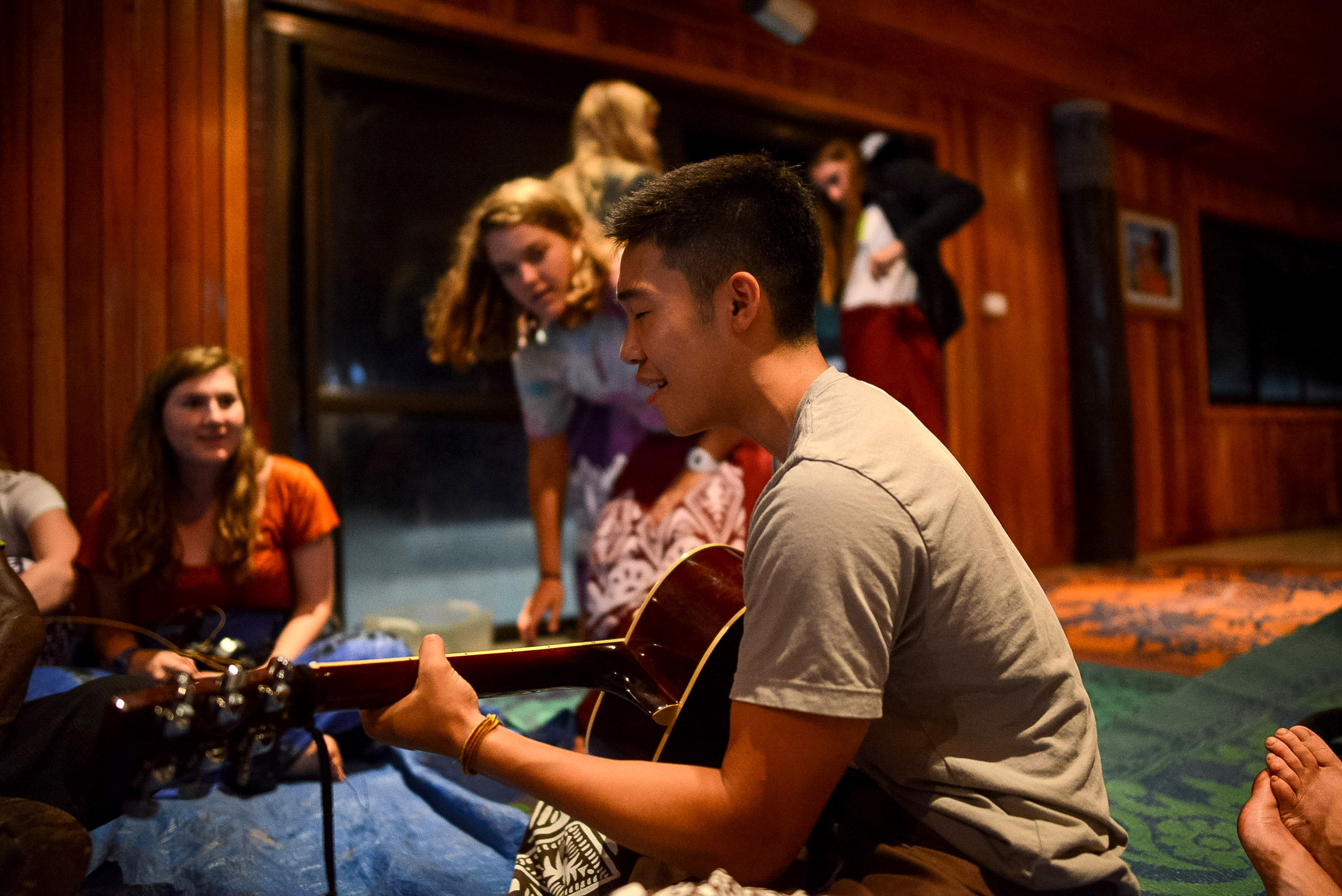 A student plays guitar while others gather around in an evening indoor hangout