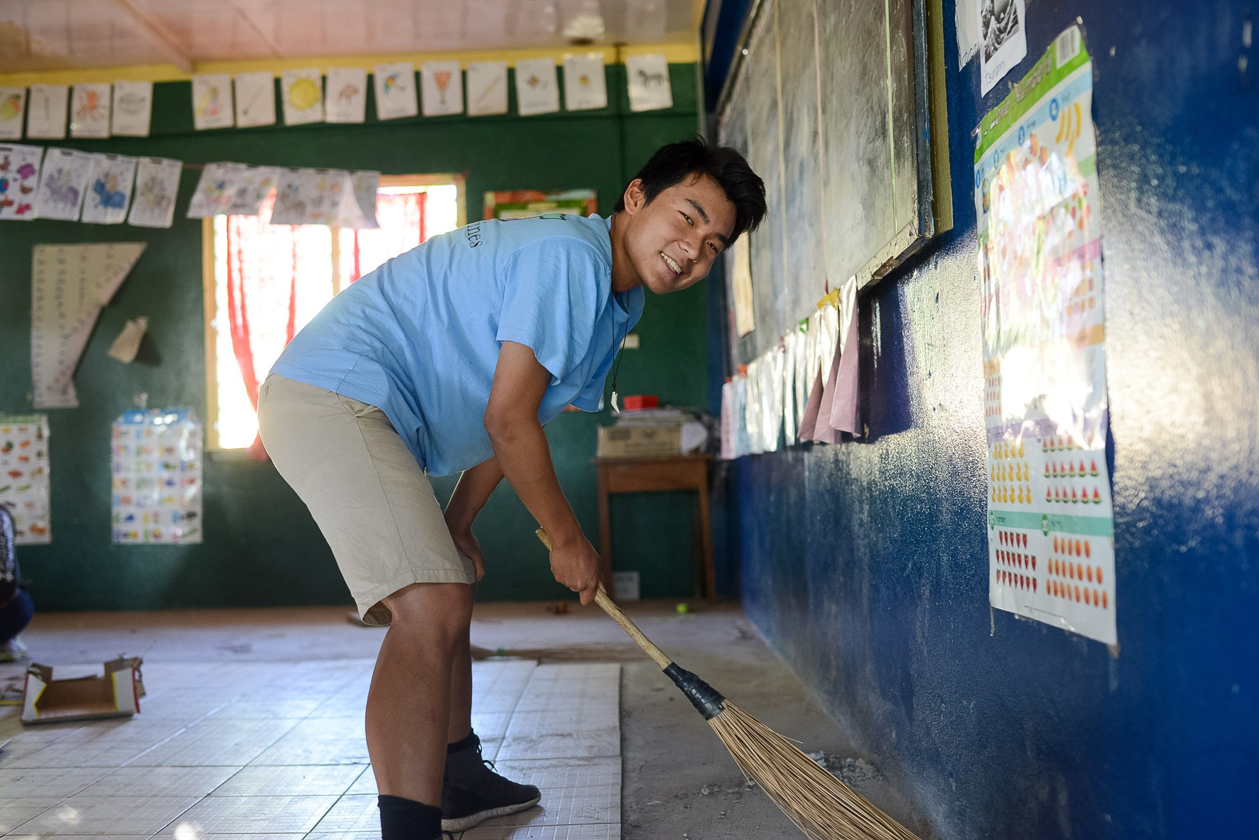 A student sweeps a classroom floor with a straw broom
