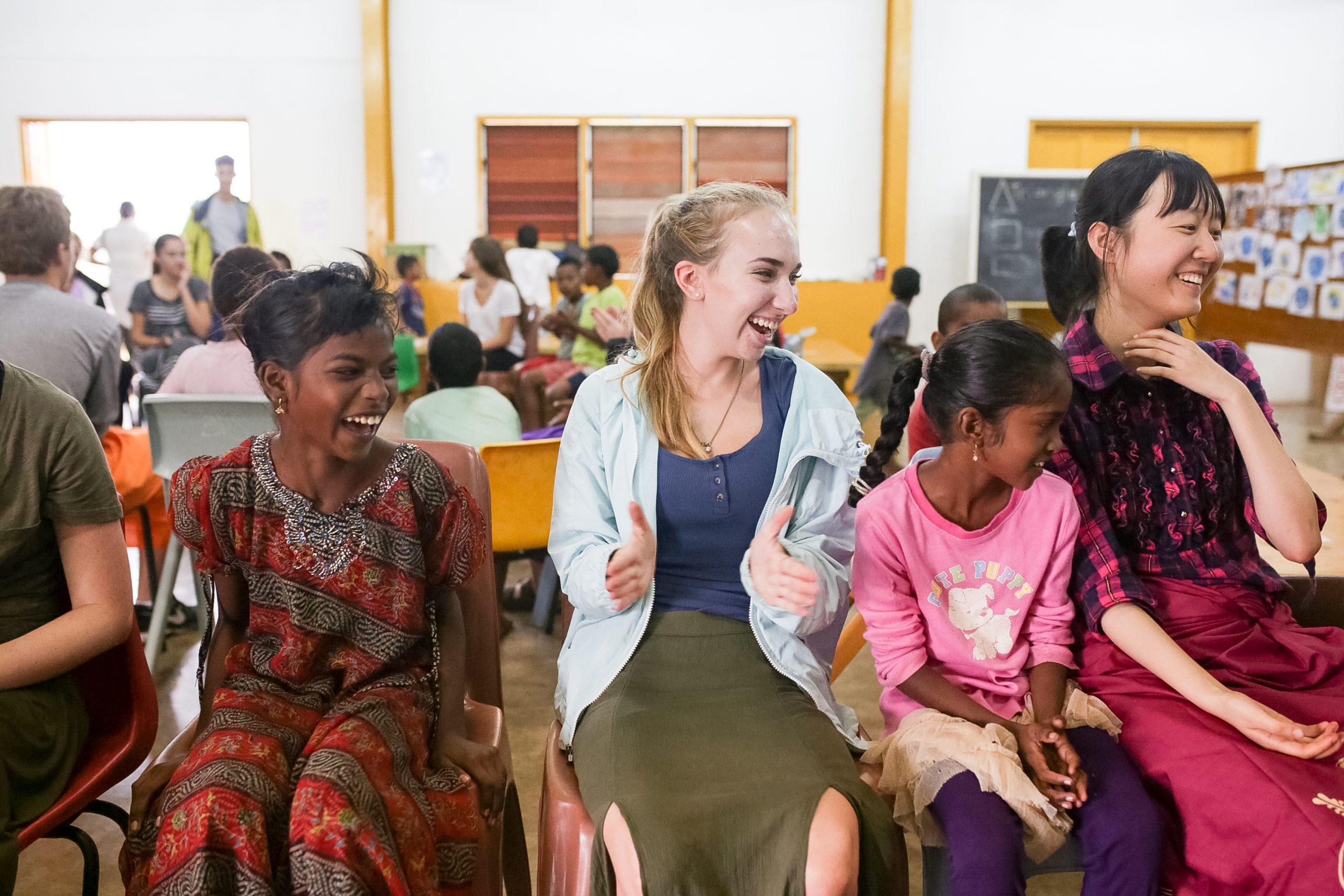 Students laugh with local children while seated in a classroom during a village visit