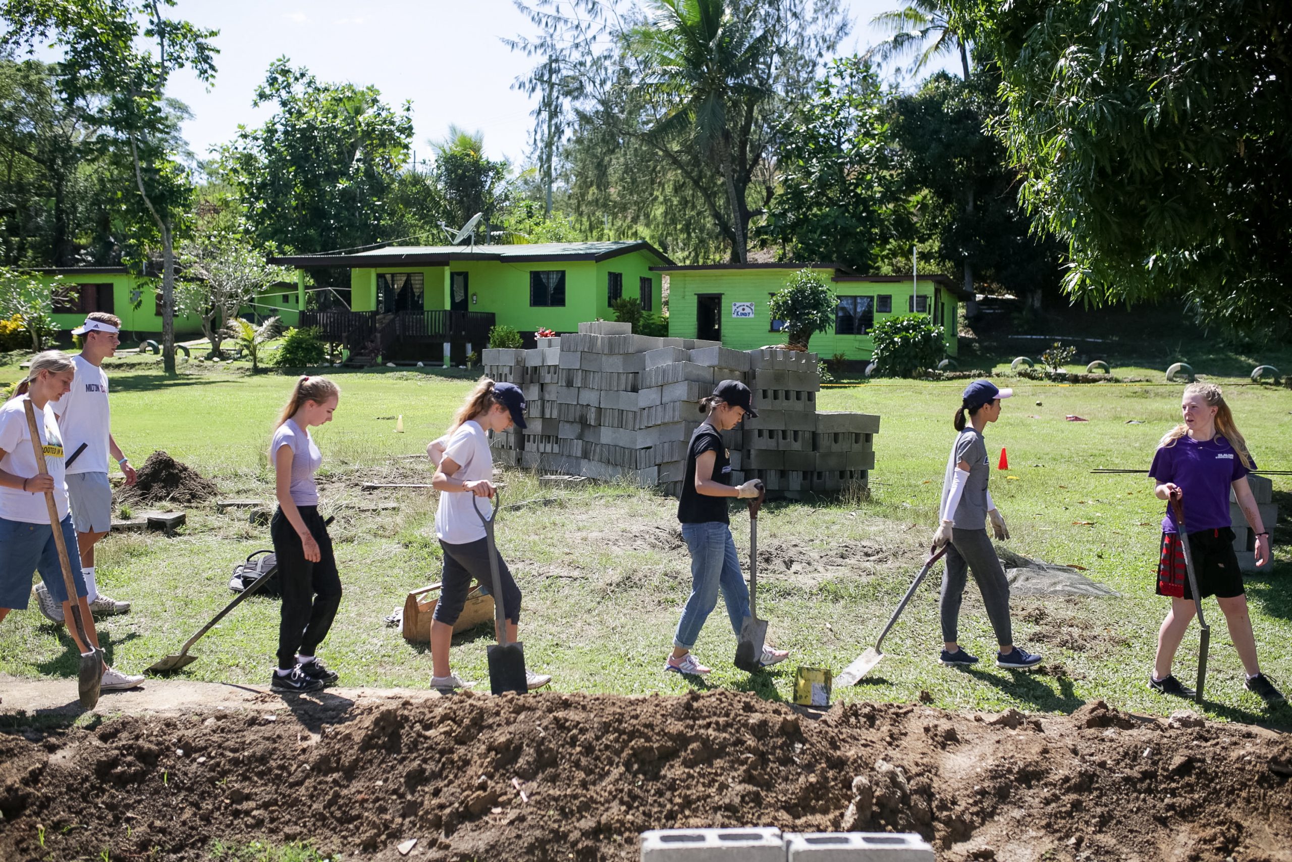 Students carry shovels and rakes across a grassy work site near stacked cinder blocks