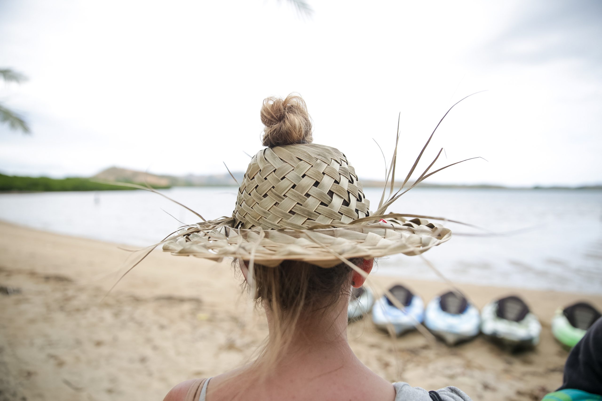 A woven straw hat seen from behind overlooks a beach lined with kayaks