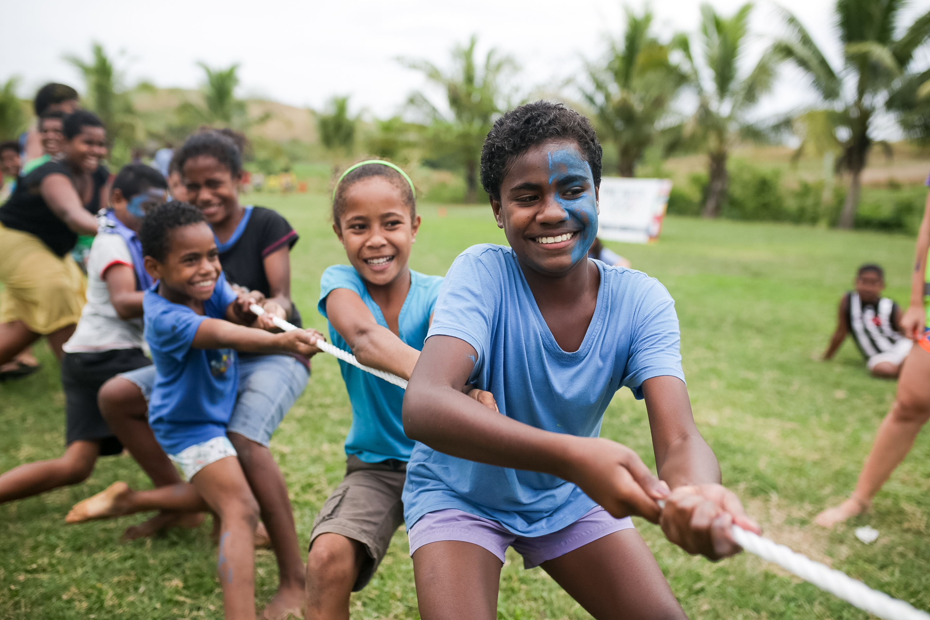 Children with painted faces play tug-of-war on a grassy field in Fiji