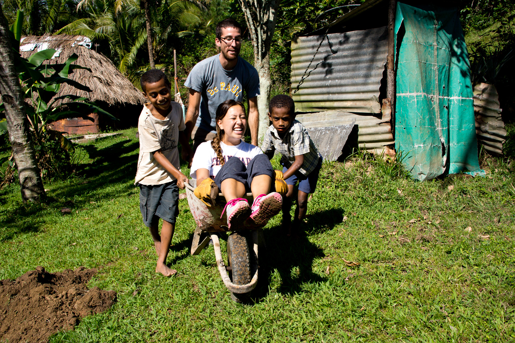 Local children push a student riding in a wheelbarrow in a village yard