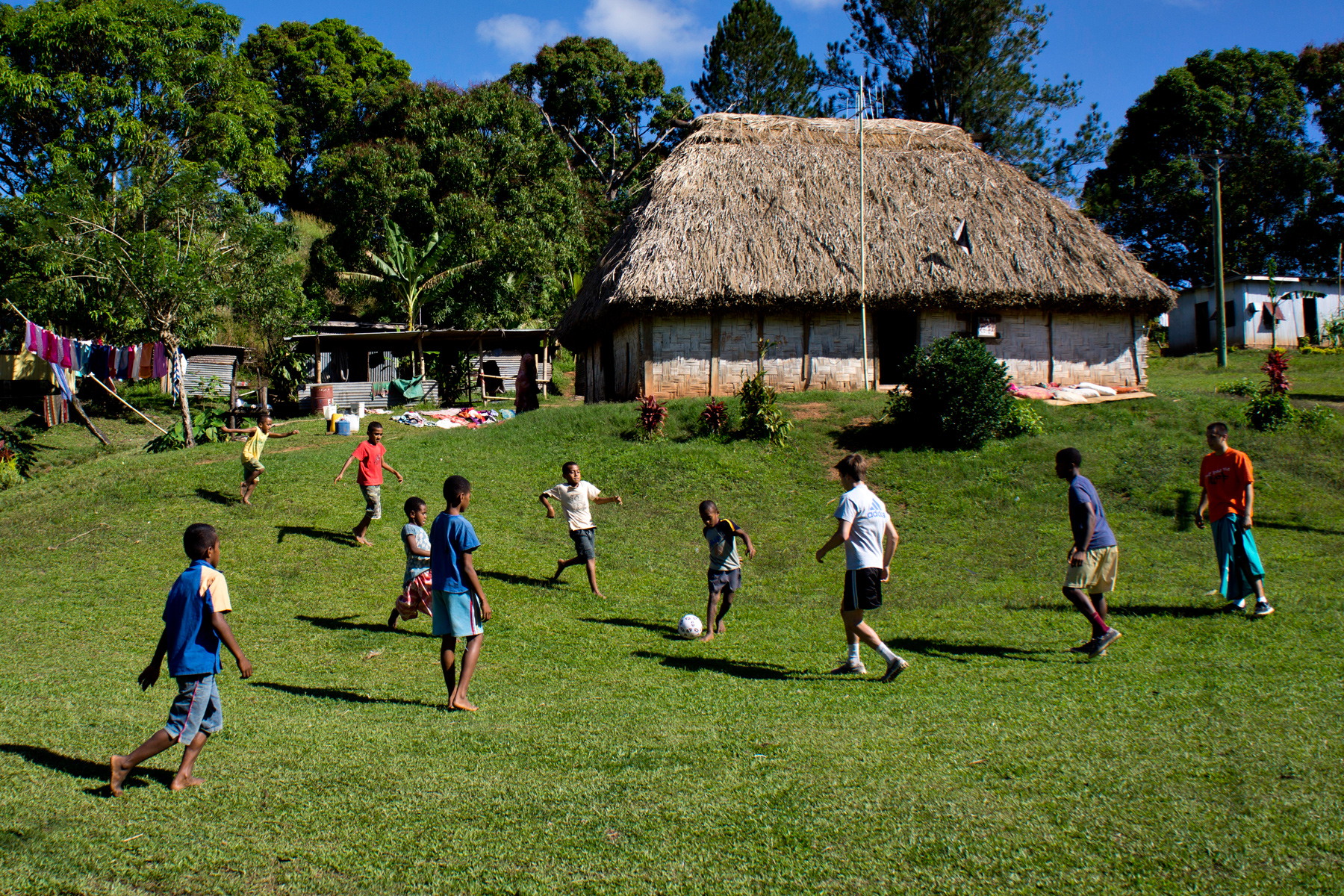Students and children play soccer on a grassy field in front of a thatched-roof Fijian bure
