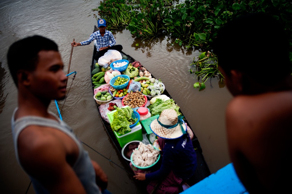 A vendor boat piled with produce navigates a river past onlookers on the bank
