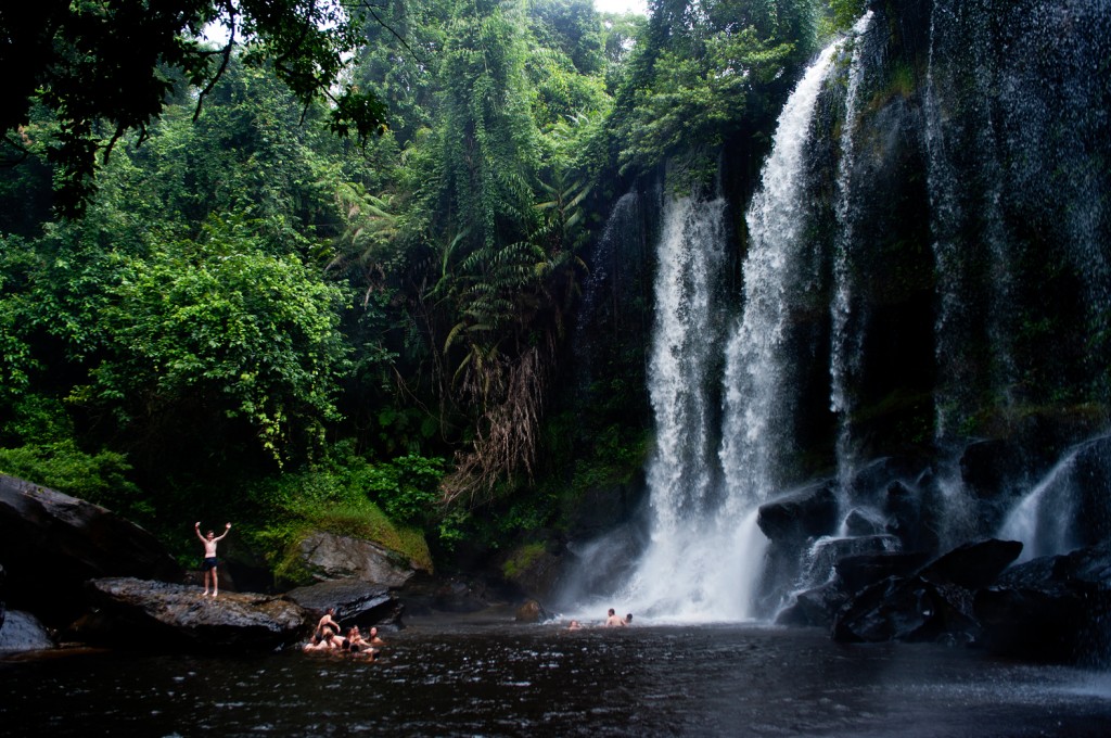 Students swim and wade in a pool beneath a jungle waterfall