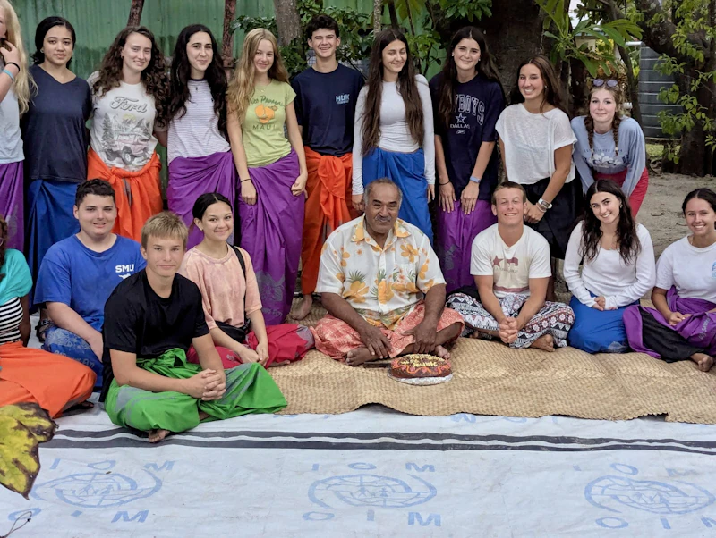 Teen travelers in brightly colored sulu sit and stand with a village elder on a woven mat during a welcome ceremony in Fiji