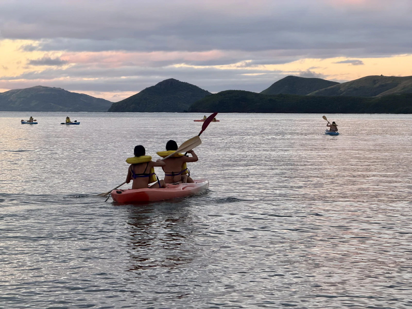 Two teen travelers paddle a red kayak on flat water at sunset, with island ridgelines and other kayaks behind them in Fiji