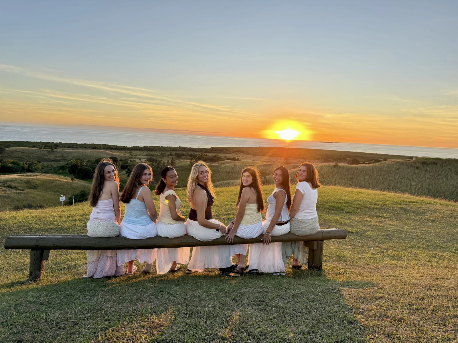 Seven teen travelers in white sit along a timber bench on a grassy headland watching the sun set over the sea in Fiji