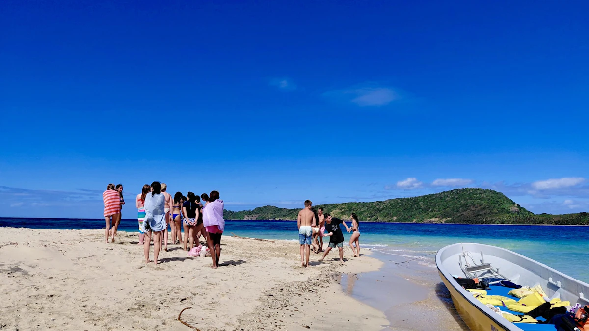 Teen travelers stand on a bright white sandbar beside a moored boat, with turquoise water and a green island behind them in Fiji