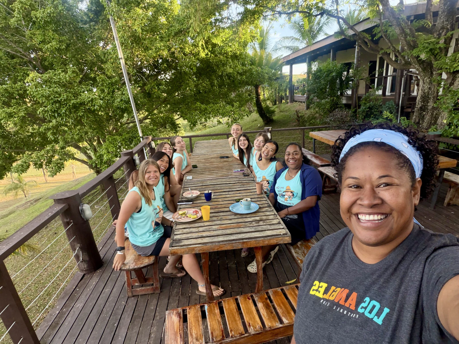 A program leader takes a selfie down a long timber table where teen travelers eat breakfast on an open deck in Fiji