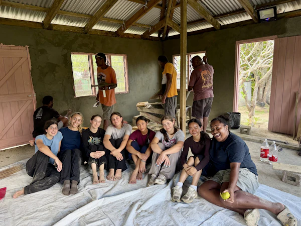 Teen travelers sit on dust sheets in a half-rendered concrete classroom while Fijian builders work on the walls behind
