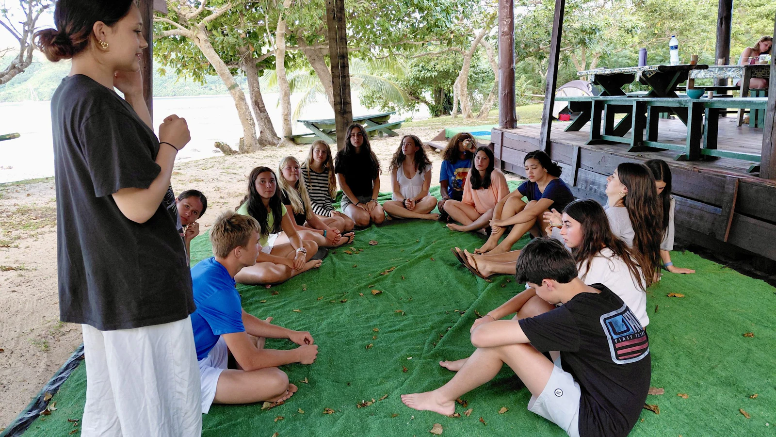 Teen travelers sit cross-legged on green mats under a shelter while a program leader briefs them, the sea behind