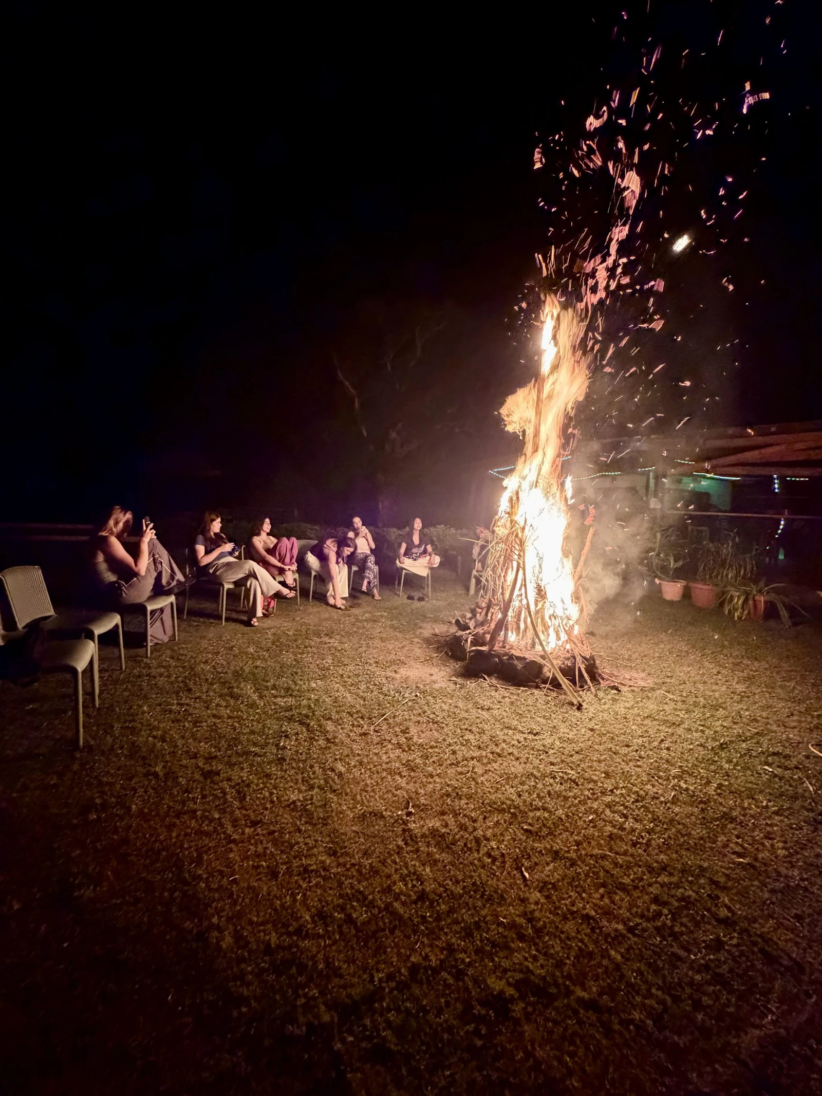 Teen travelers sit on chairs in a semicircle around a tall bonfire throwing sparks into the night sky in Fiji