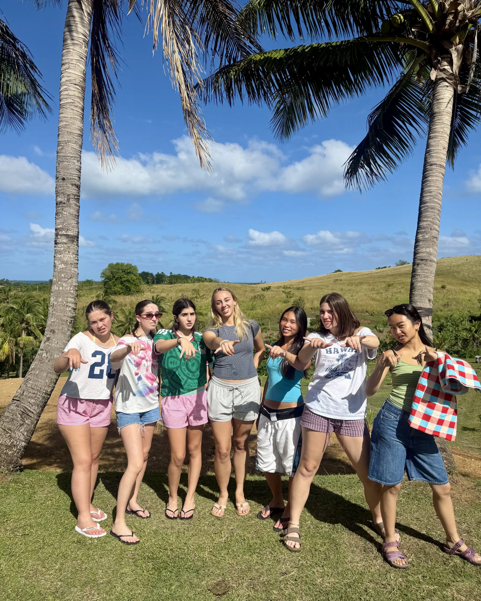 Seven teen travelers stand pointing at the camera between two coconut palms with green hills behind them in Fiji