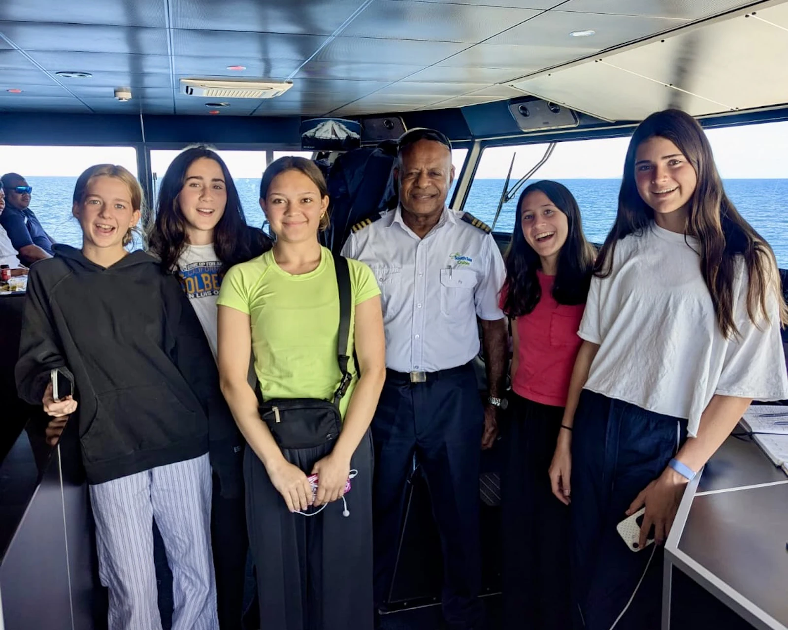 Five teen travelers stand with a Fijian ferry captain in a white uniform shirt on the bridge of a boat at sea