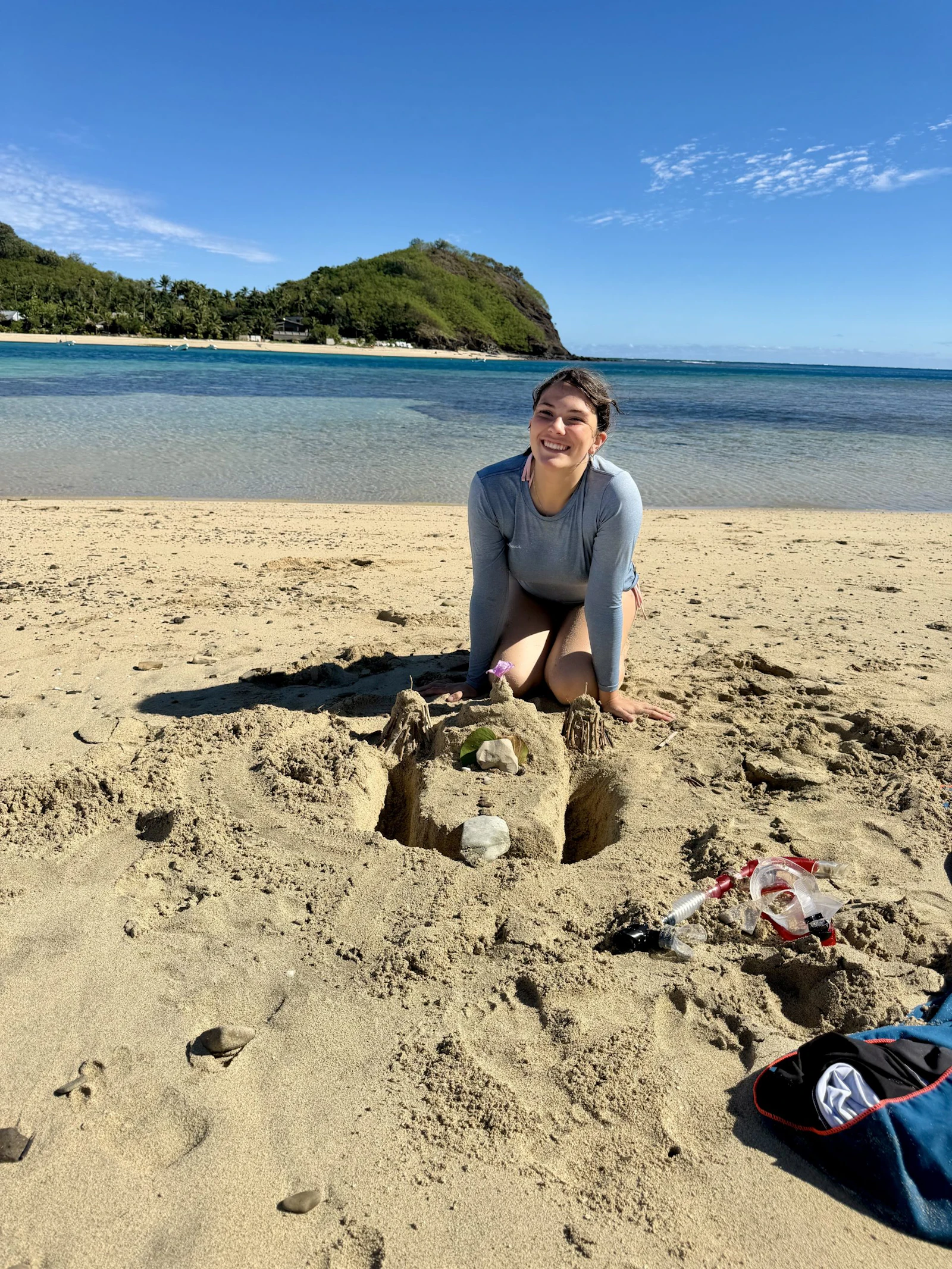 A teen traveler kneels beside a large sea turtle sculpted in wet sand on a Fijian beach, snorkel gear beside her