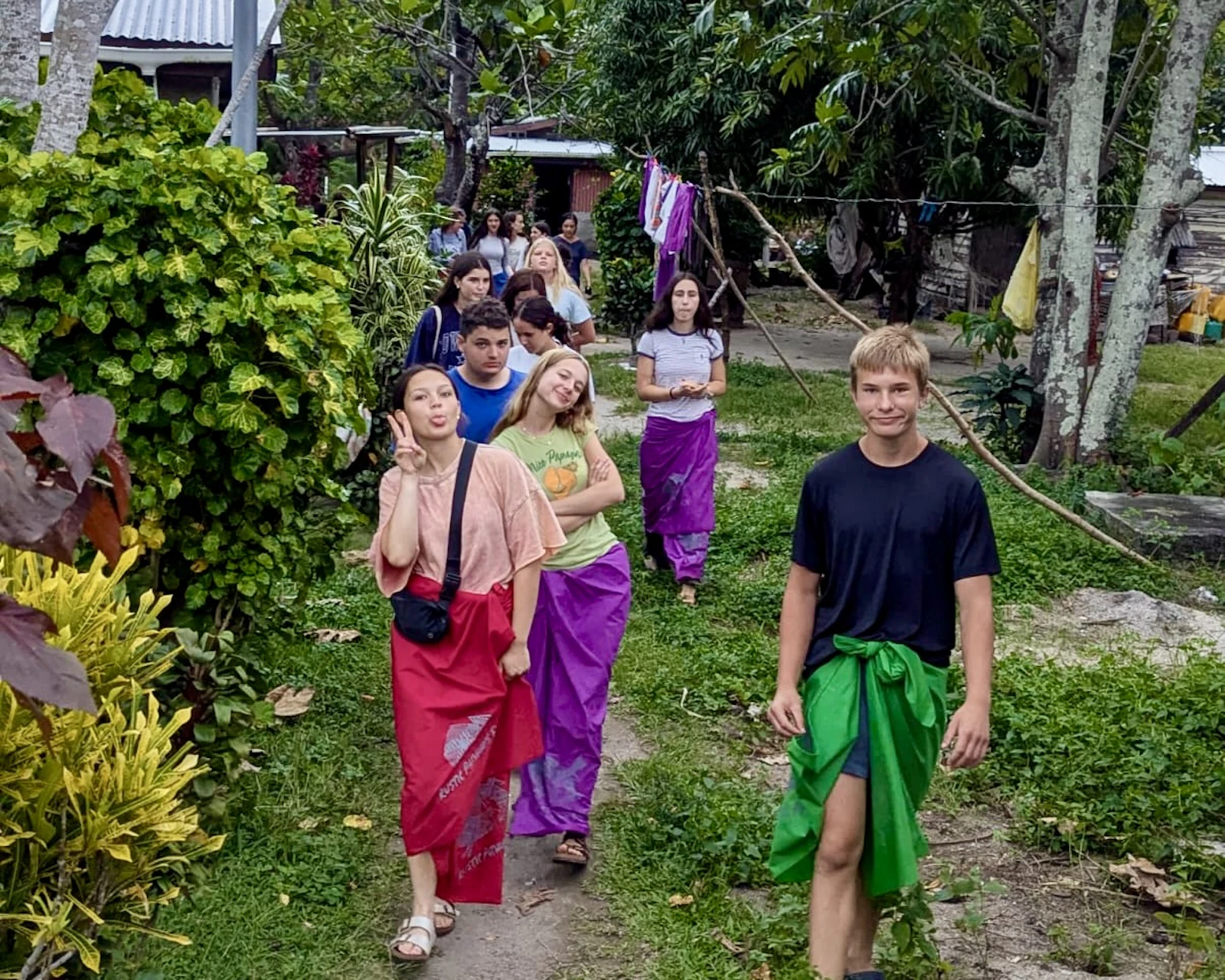 Teen travelers in colorful sulu wraps walk a dirt path through a Fijian village, houses and washing lines behind them